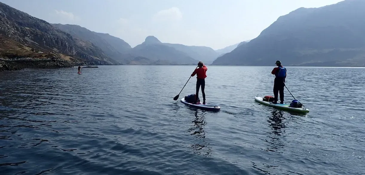 Mae dau berson yn padlfyrddio ar lyn gyda mynyddoedd yn y cefndir.