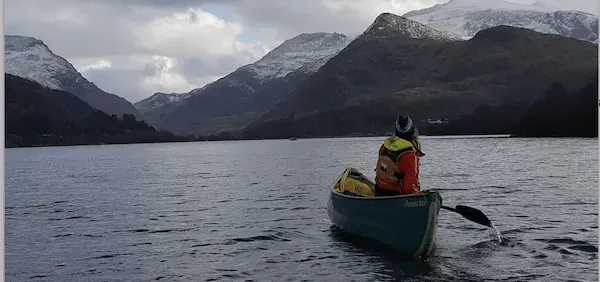 Mae person mewn canŵ ar lyn gyda mynyddoedd yn y cefndir.