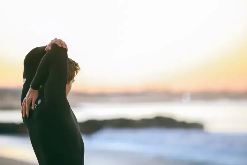 A man in a wetsuit is stretching his arms on the beach.