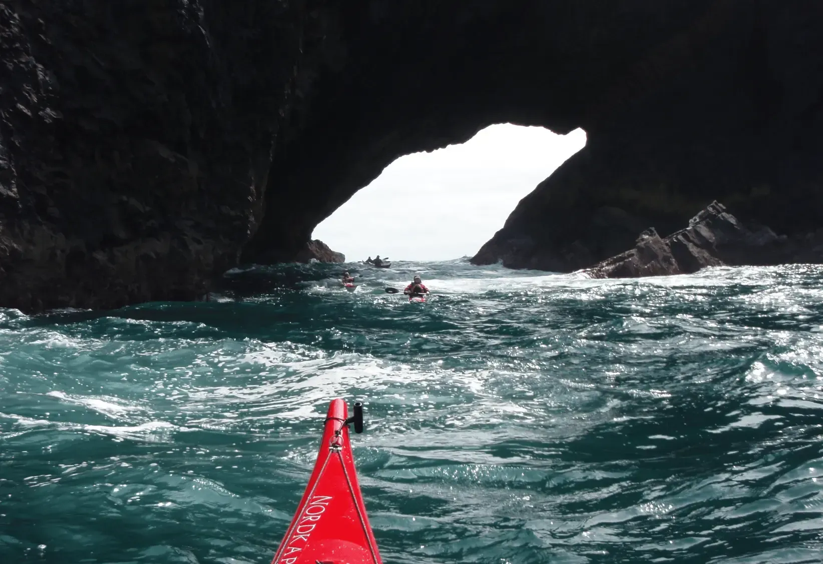 Mae caiac coch yn y dwr ger ogof