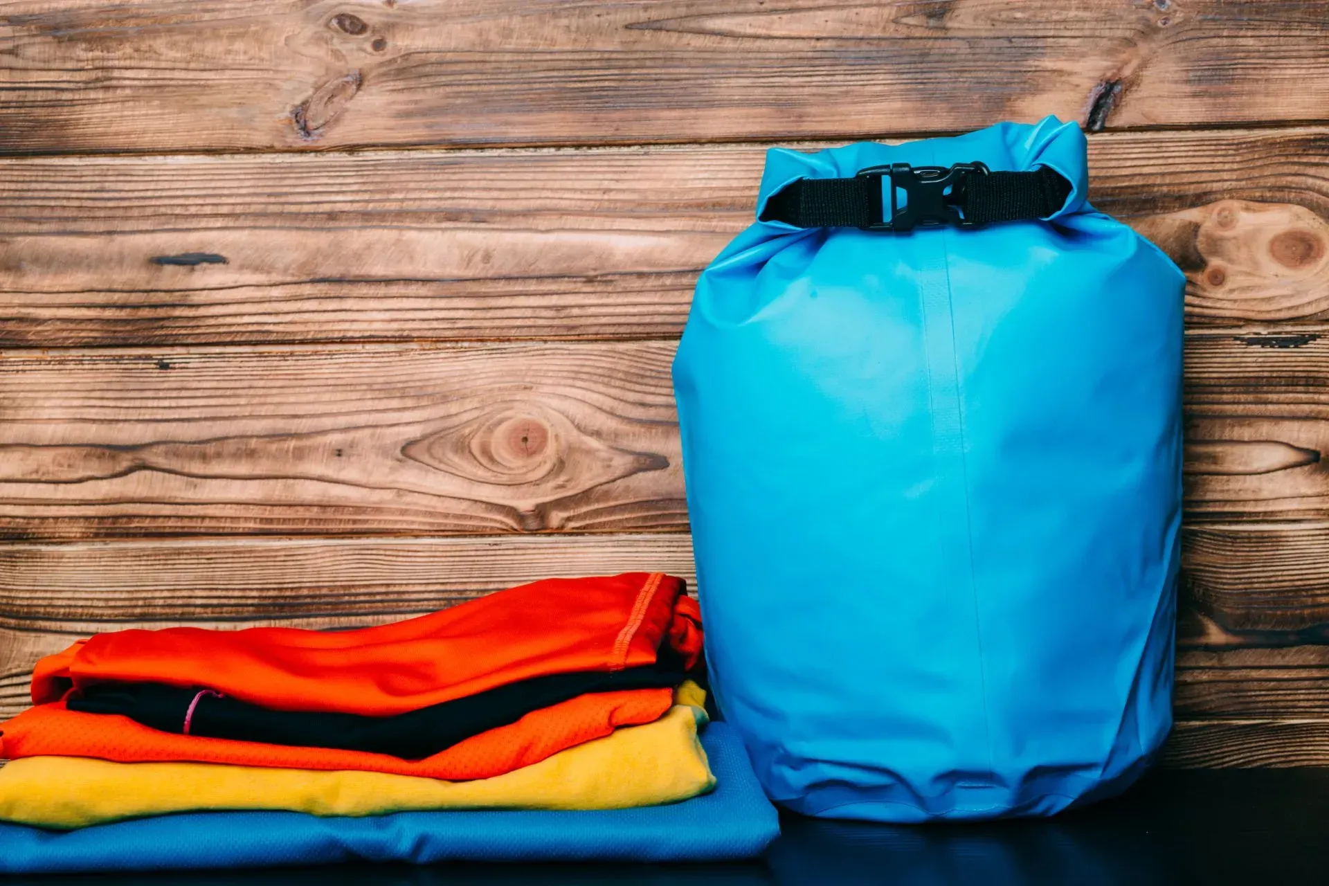 A blue dry bag is sitting next to a pile of clothes on a wooden table.