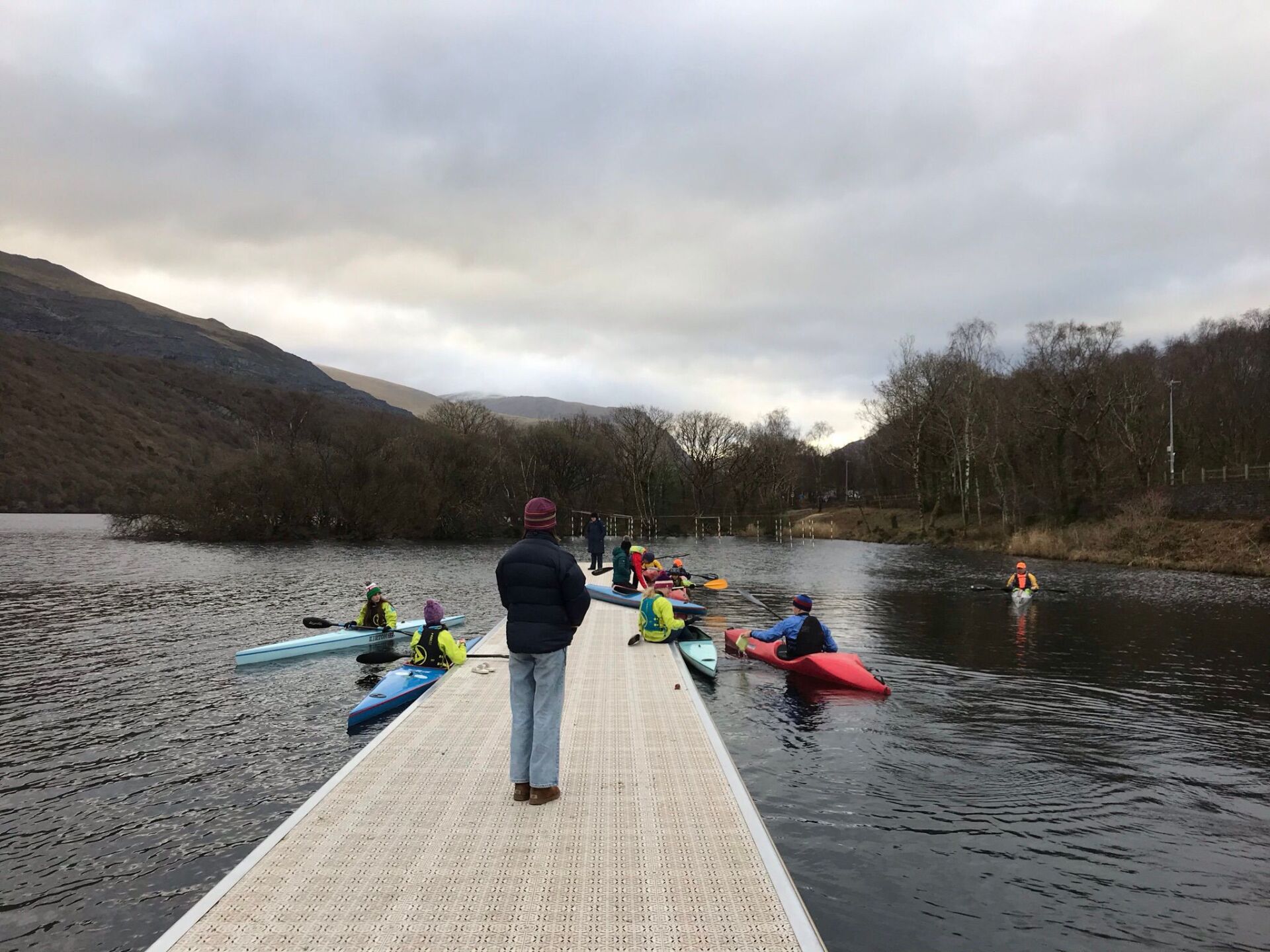 A person is standing on a dock overlooking a lake with people in kayaks.