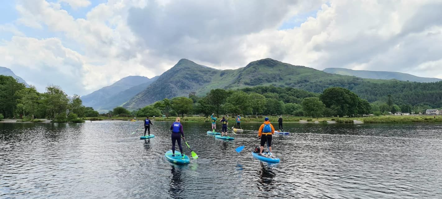 Mae criw o bobl yn padlfyrddio ar lyn gyda mynyddoedd yn y cefndir.