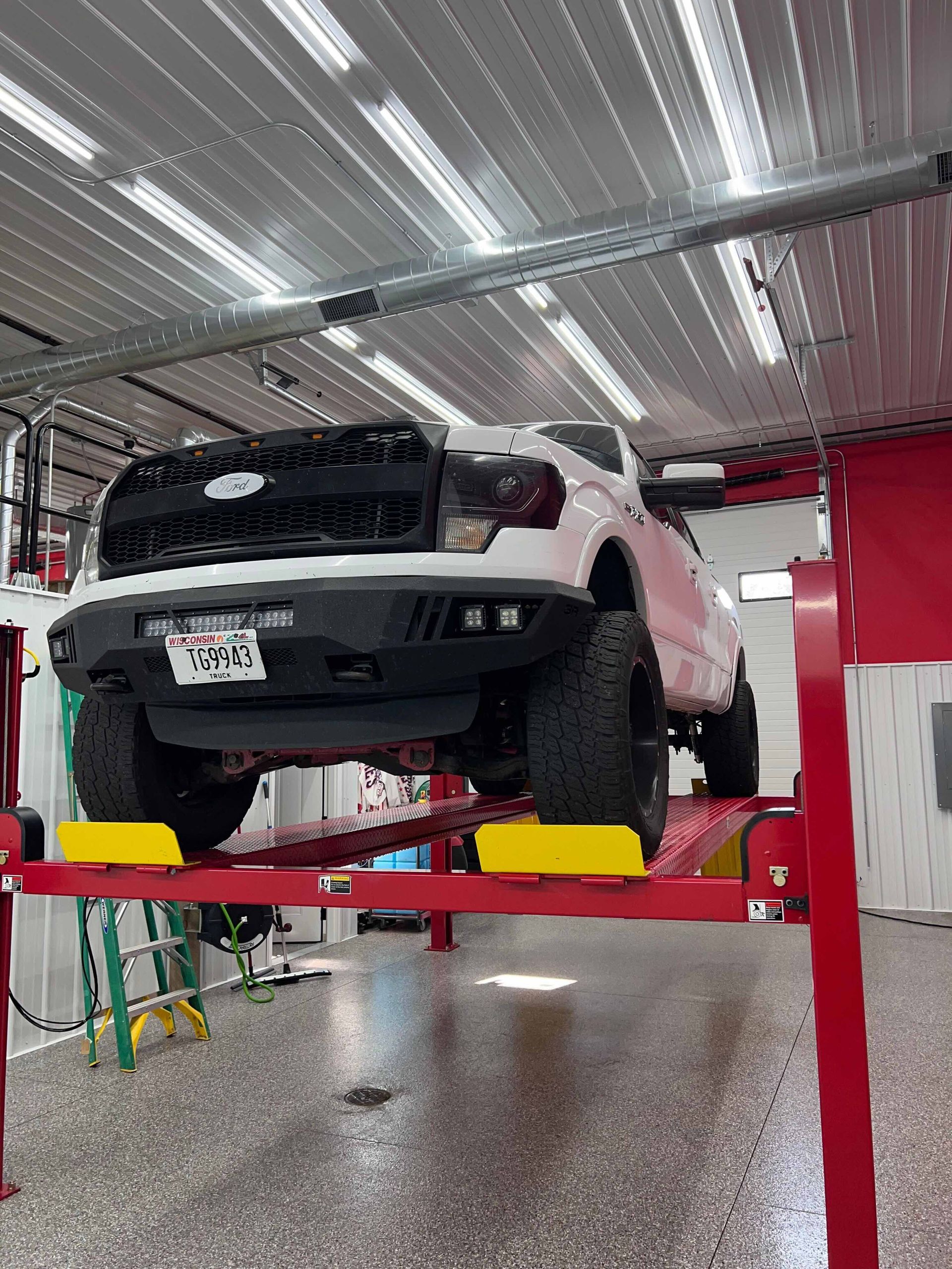 White pickup truck on a red hydraulic lift in a garage with a speckled floor and white walls.