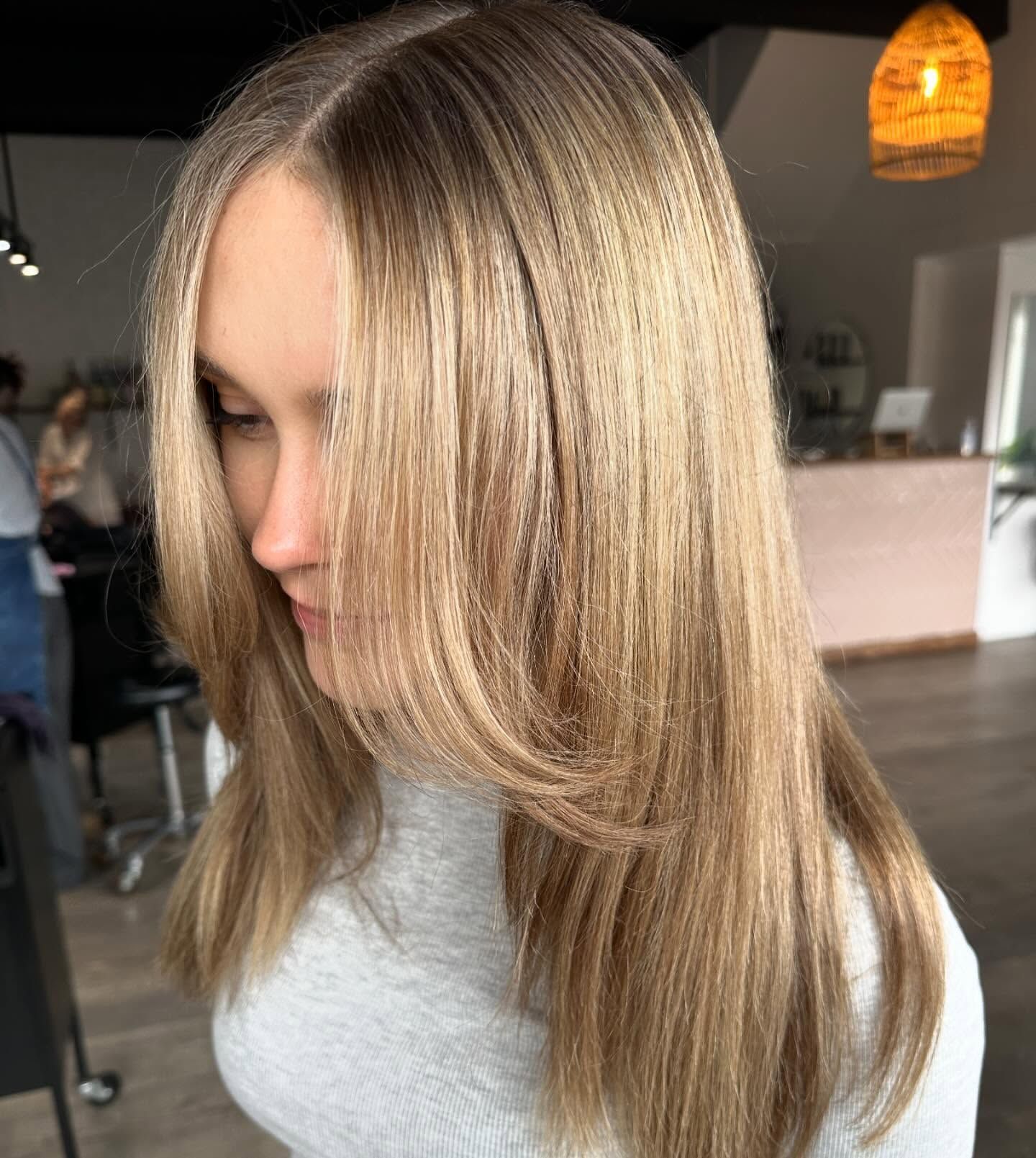 Woman with long, layered, light brown hair. She wears a gray sweater, in a salon setting.
