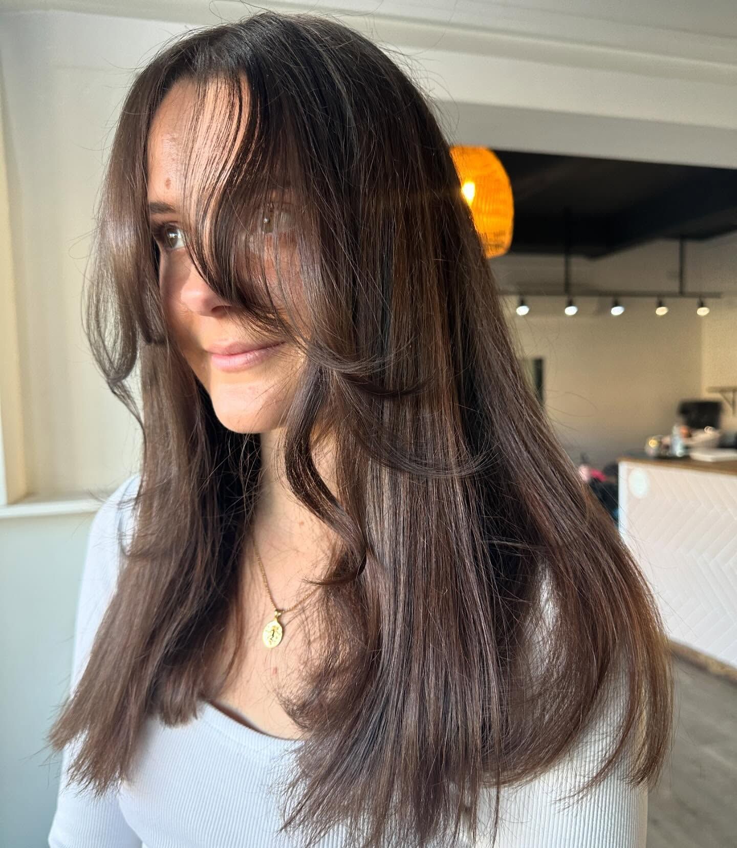 Woman with long, layered brown hair and face-framing bangs; inside a salon, wearing white top, necklace.