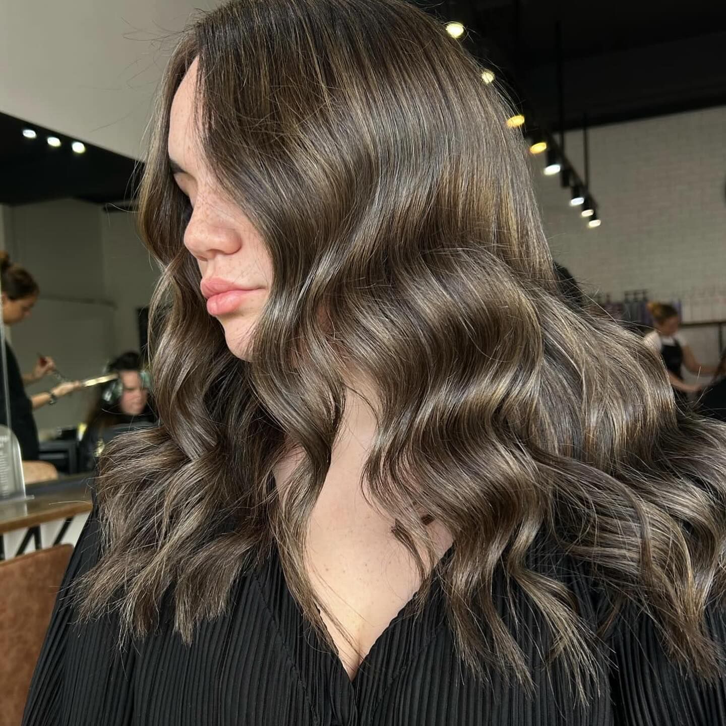 Woman with wavy, dark brown hair. She wears a black top, and is in a salon setting.