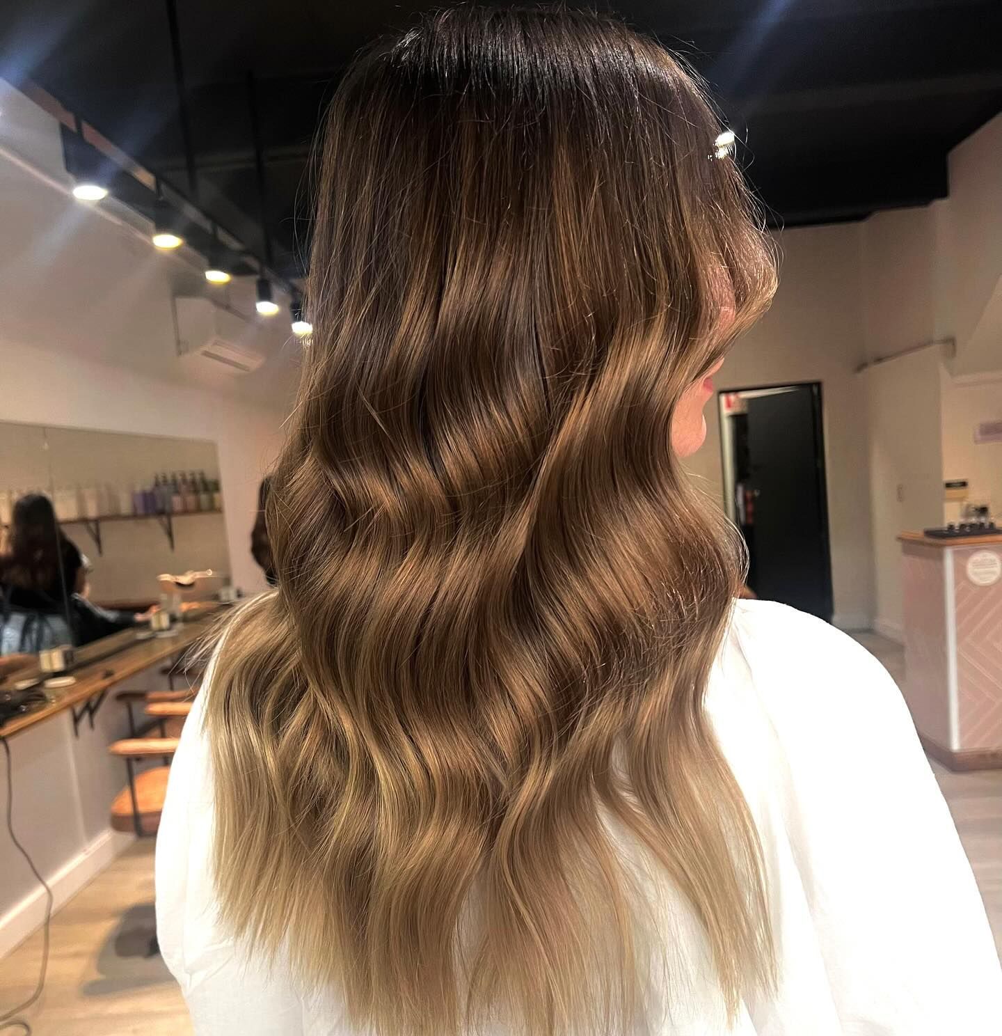 Woman with wavy, brunette hair, ombre style. Light brown at the ends. Inside a salon.