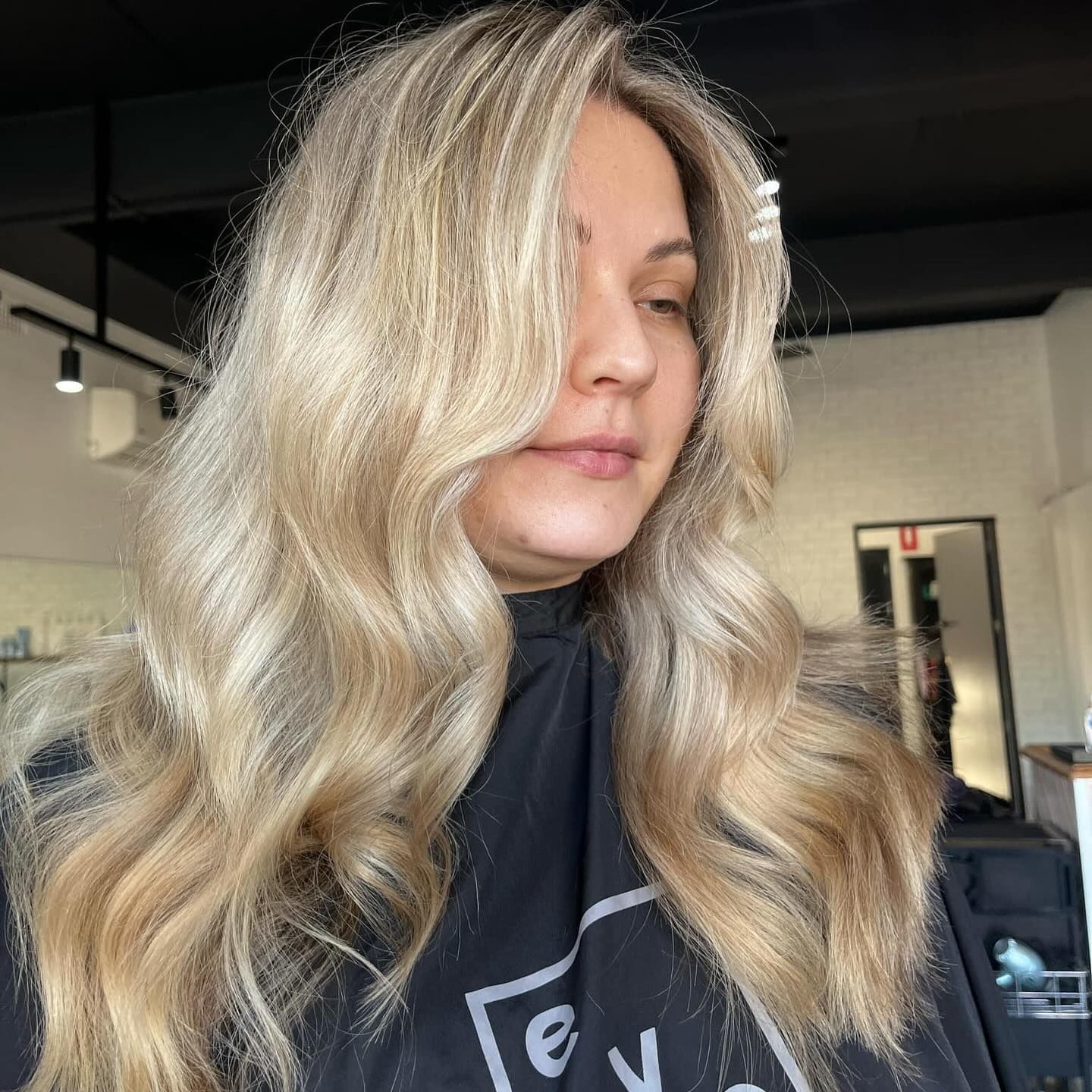 Woman with long, wavy blonde hair in a salon setting, wearing a black styling cape.