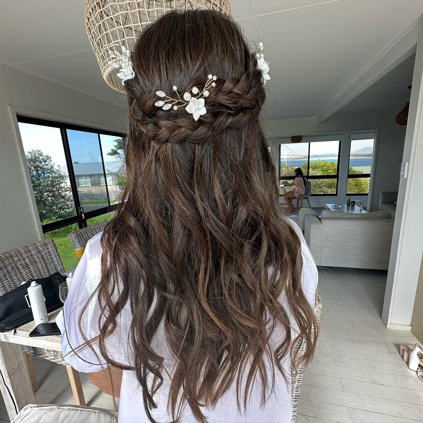Woman with wavy brown hair styled in a half-updo with floral hair accessories, in a bright room with a view.