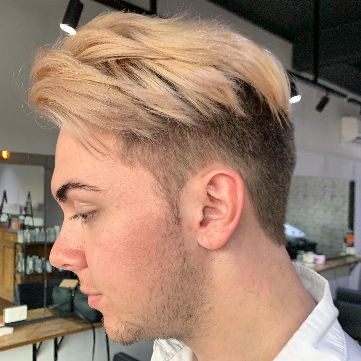 Man with light blonde hair, undercut style. He is in a salon, facing left, with a neutral expression.