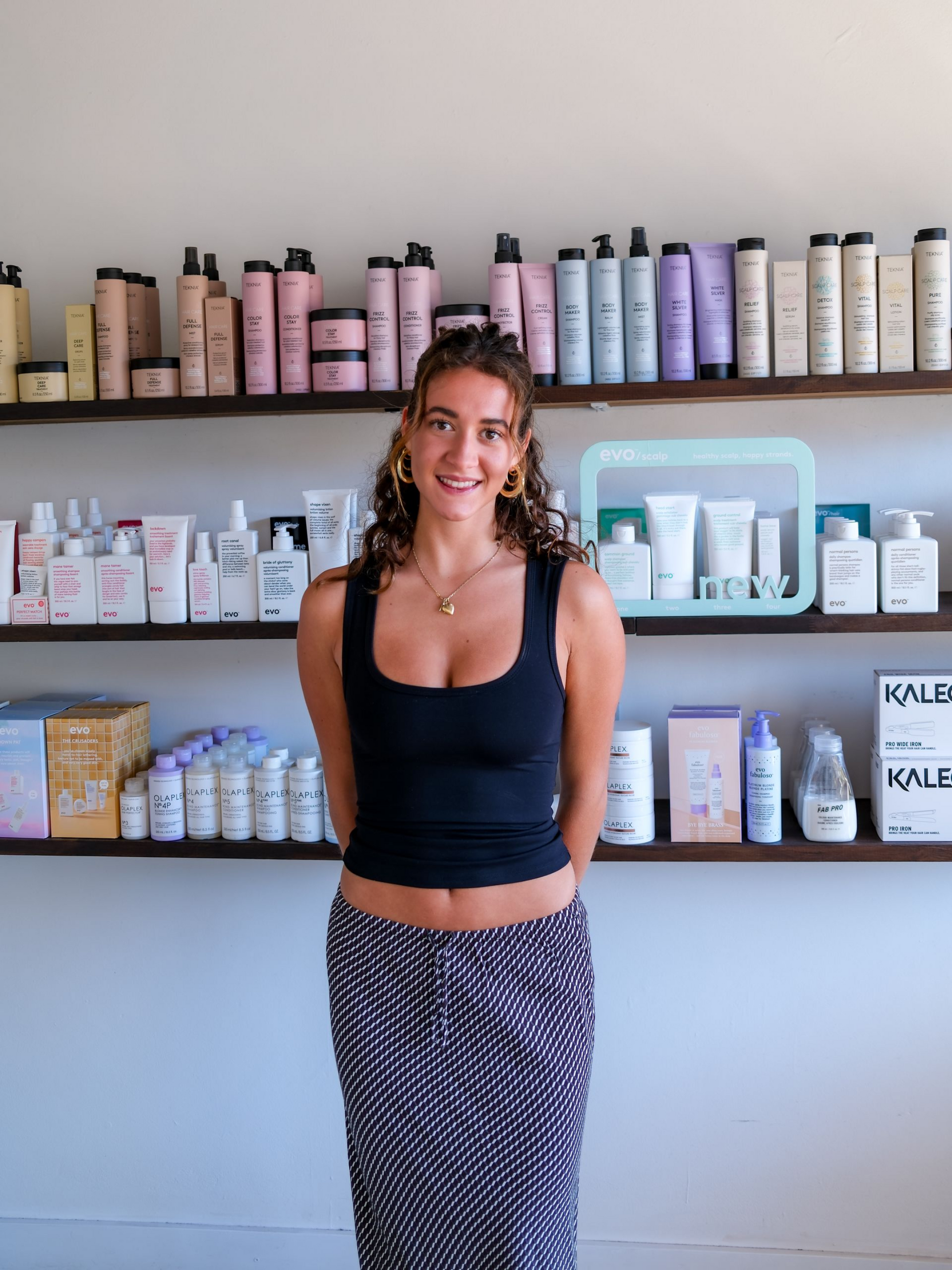 Woman in black tank top smiles in front of shelves filled with beauty products.