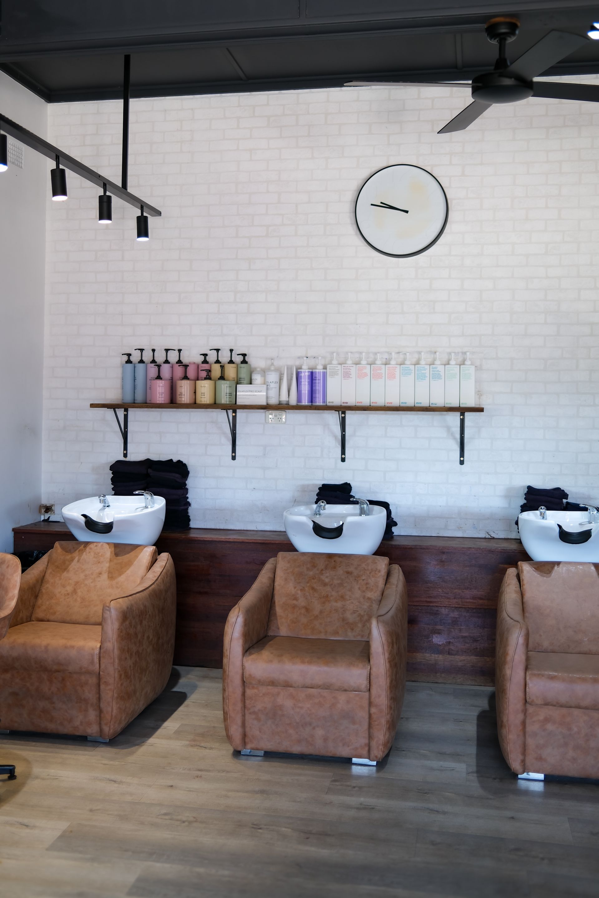 Salon with three tan chairs, wash basins, and a shelf of hair products against a white brick wall.