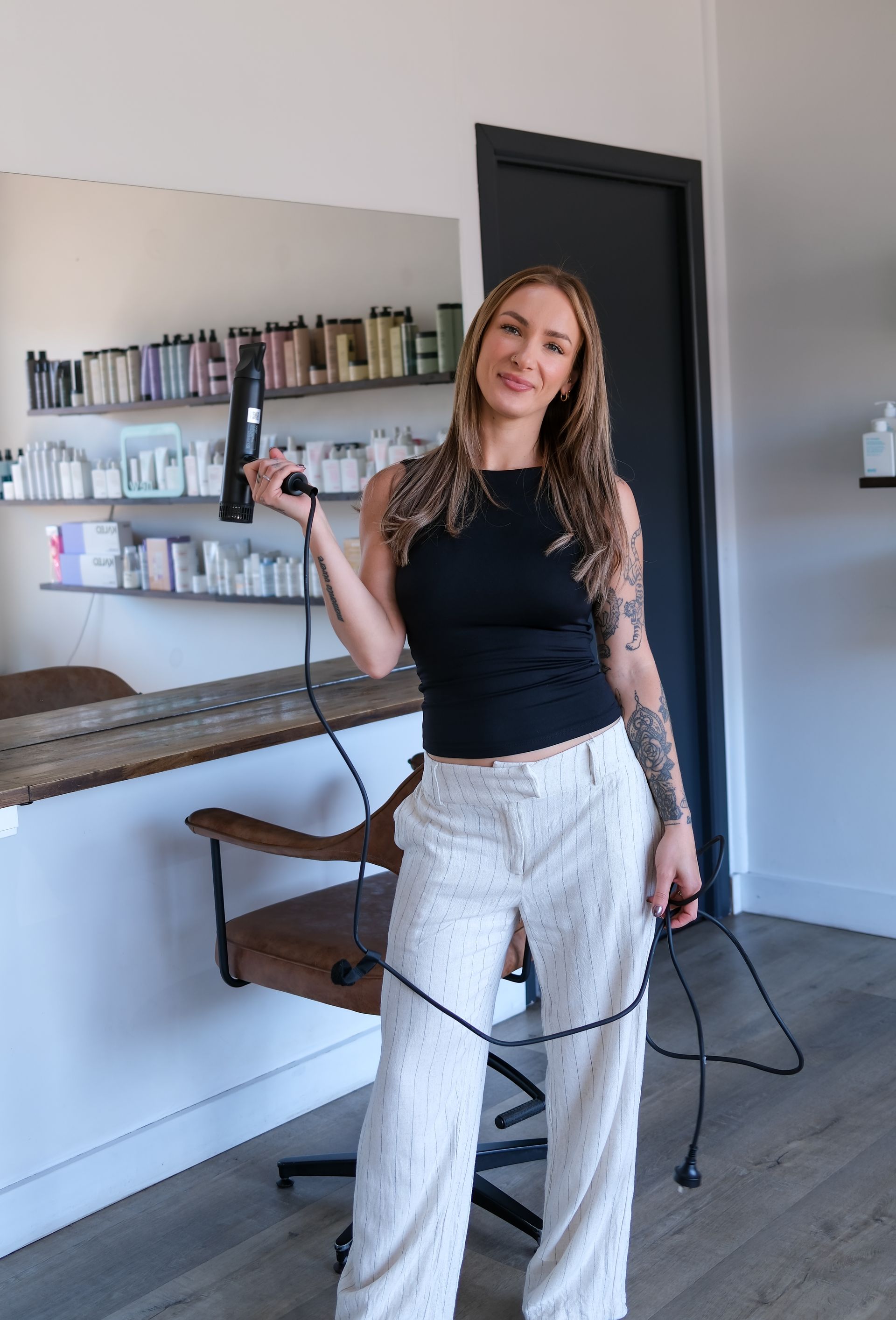 Woman in salon holding hairdryer, smiling. Wearing black top, white pants, with arm tattoo.