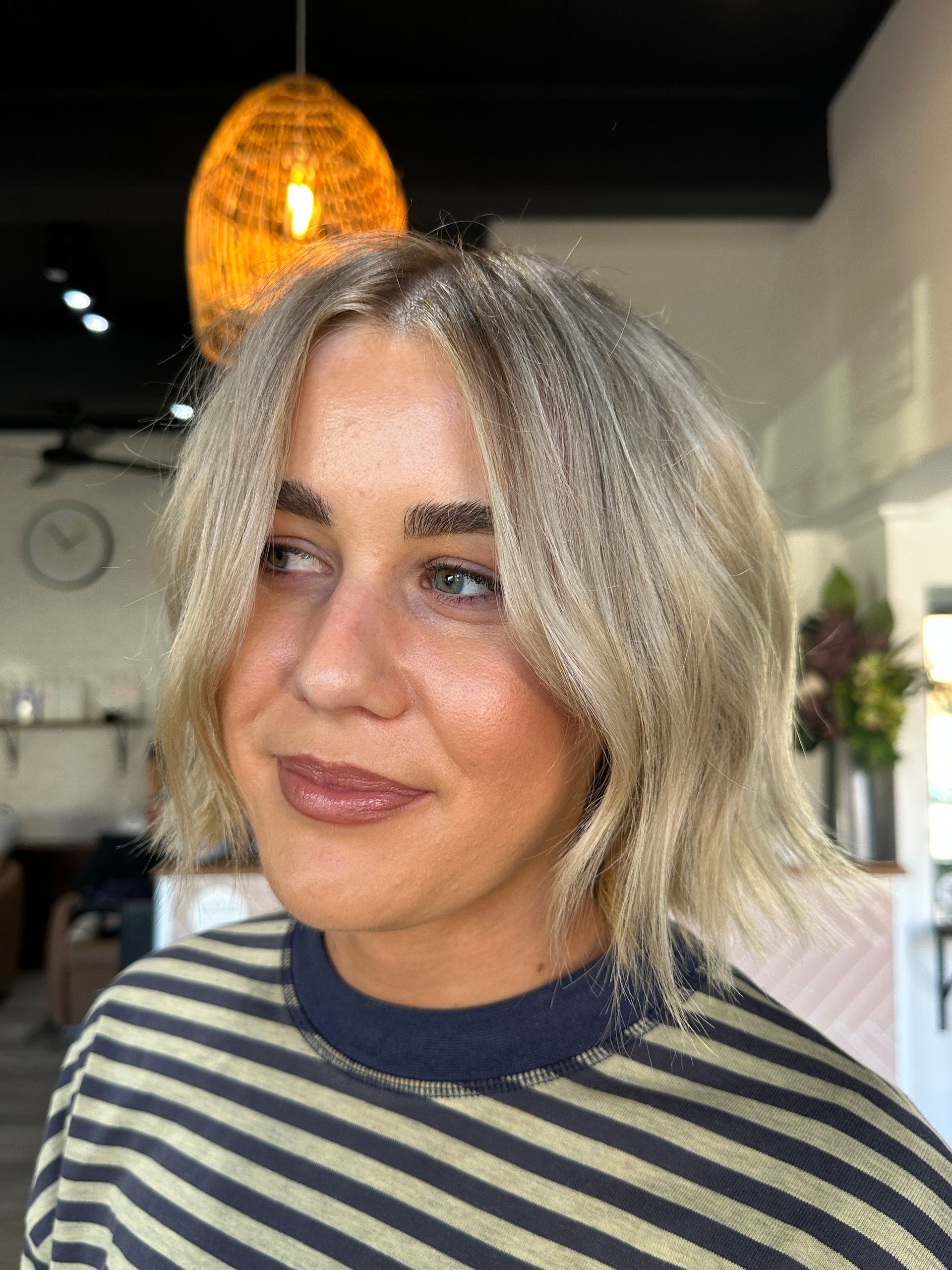Woman with a short, layered blonde hairstyle. Wearing a striped shirt, she is smiling inside a salon.