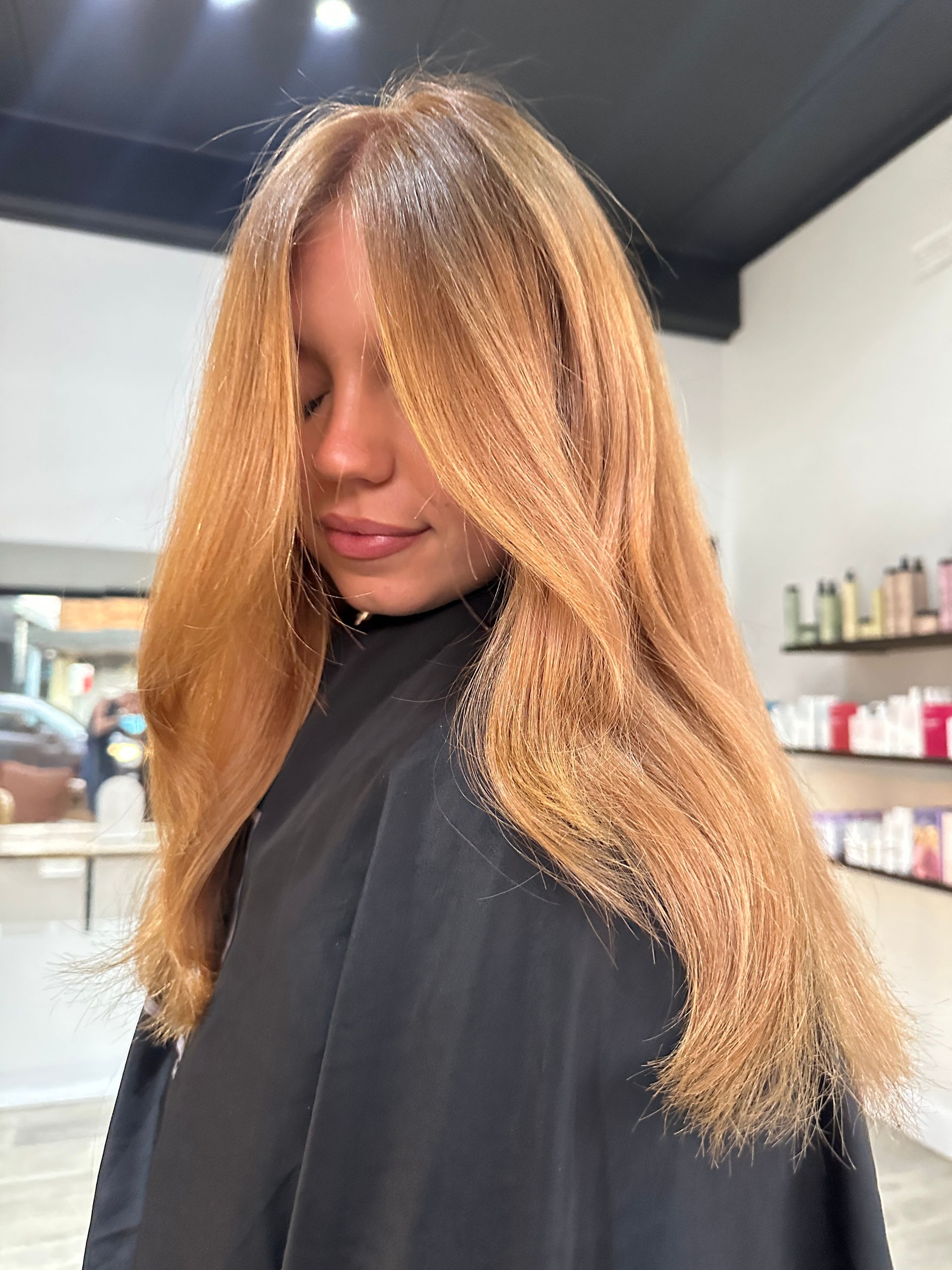 Woman with long, reddish-blonde hair in a salon, wearing a black cape.