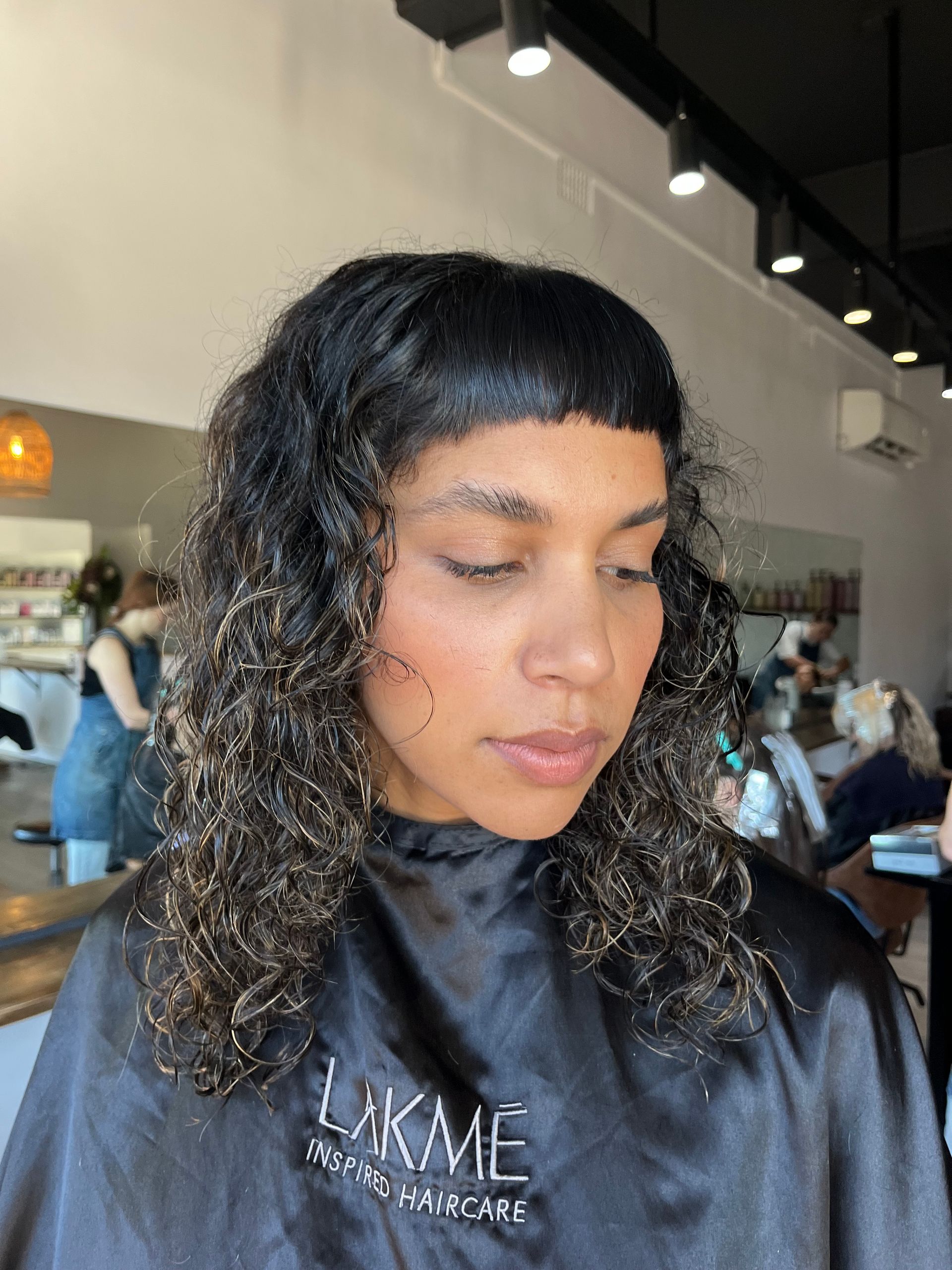 Woman with dark, curly hair in a salon, wearing a black cape. Bangs, neutral expression.