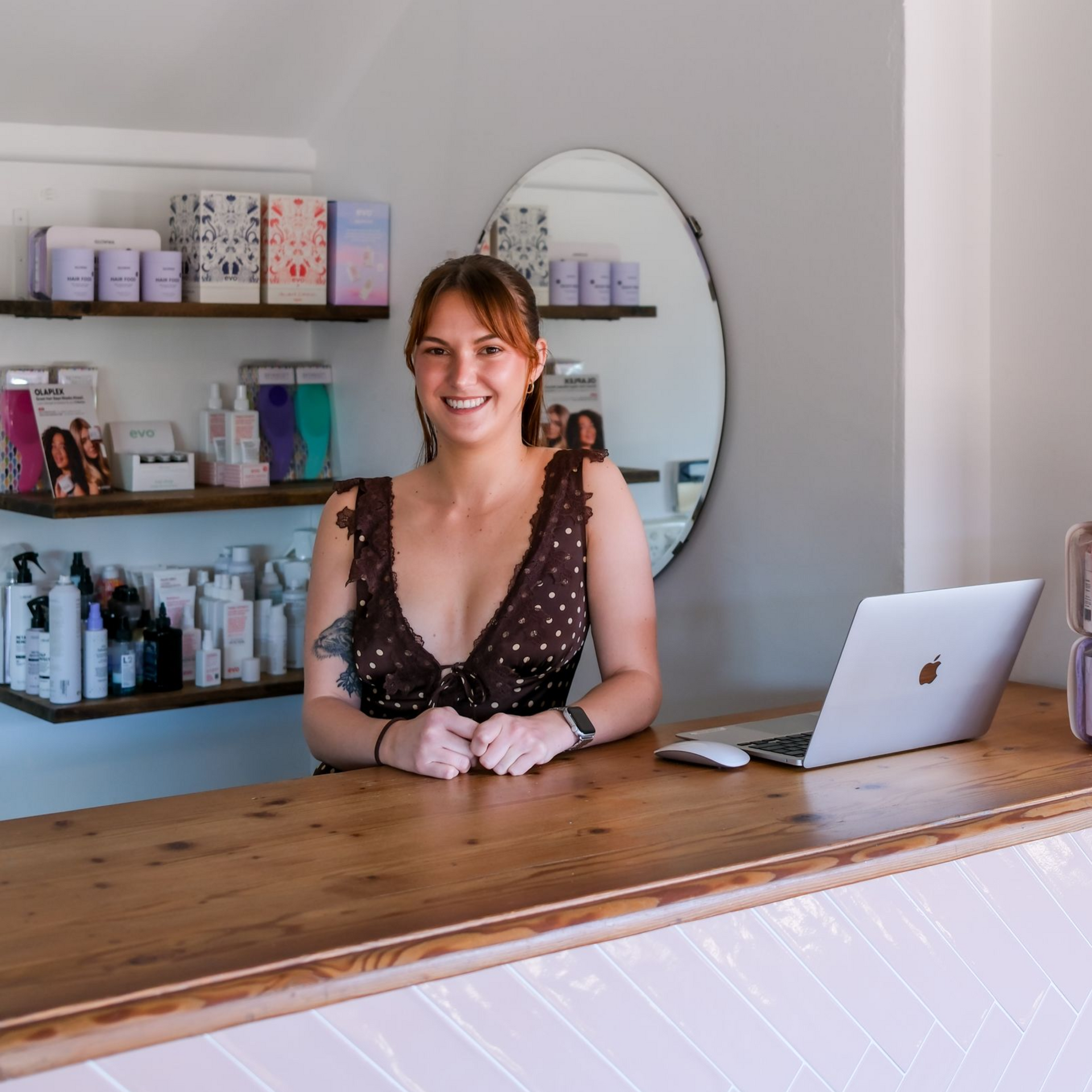 Woman at salon counter with laptop and products, smiling.