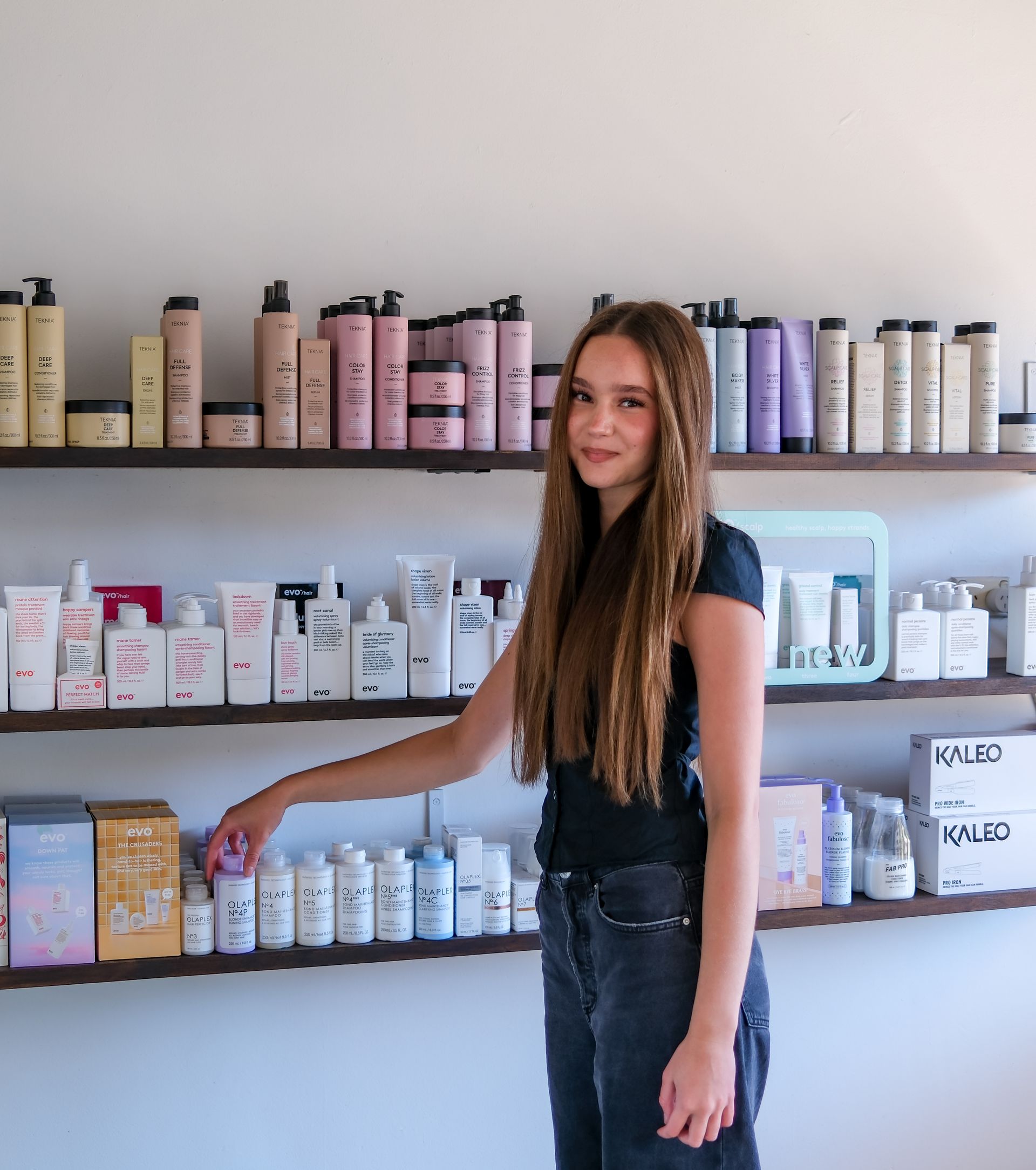 Woman pointing at a product on a shelf of beauty products; white bottles and pink packaging.