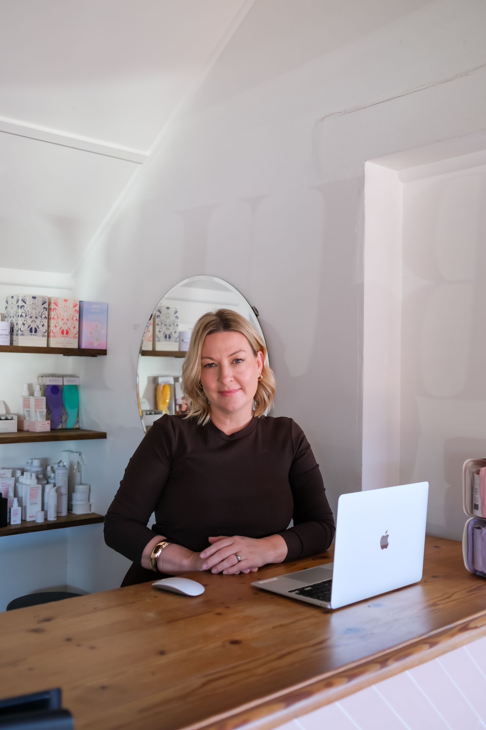 Woman at a wooden desk with a laptop, smiling. Retail setting with shelves of products.