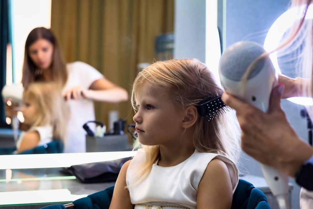 Girl getting hair styled in a salon, with a stylist using a hairdryer and another combing hair in the mirror's reflection.