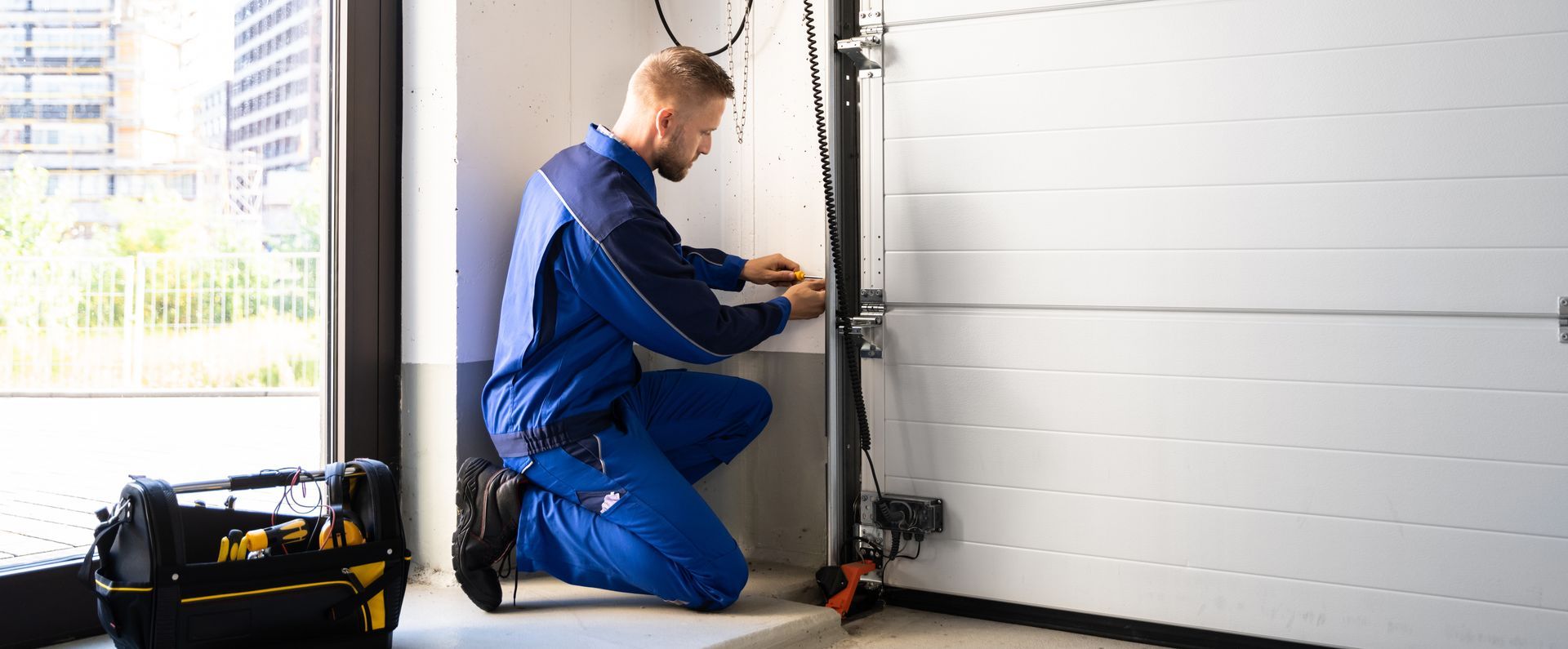 Technician in blue coveralls repairing a garage door mechanism beside a tool bag.