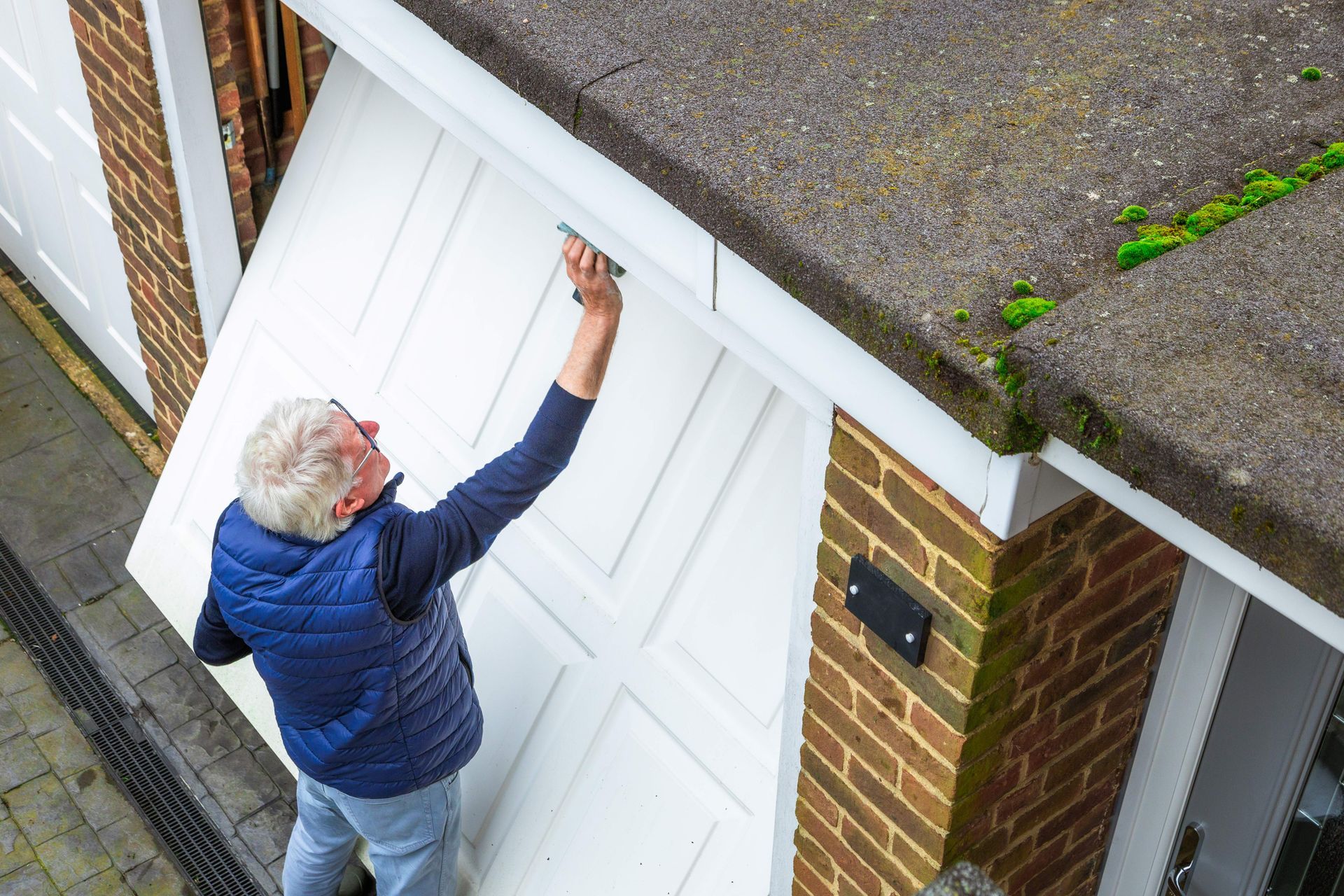 Older adult inspecting white garage door with tool near brick wall and moss-covered roof.