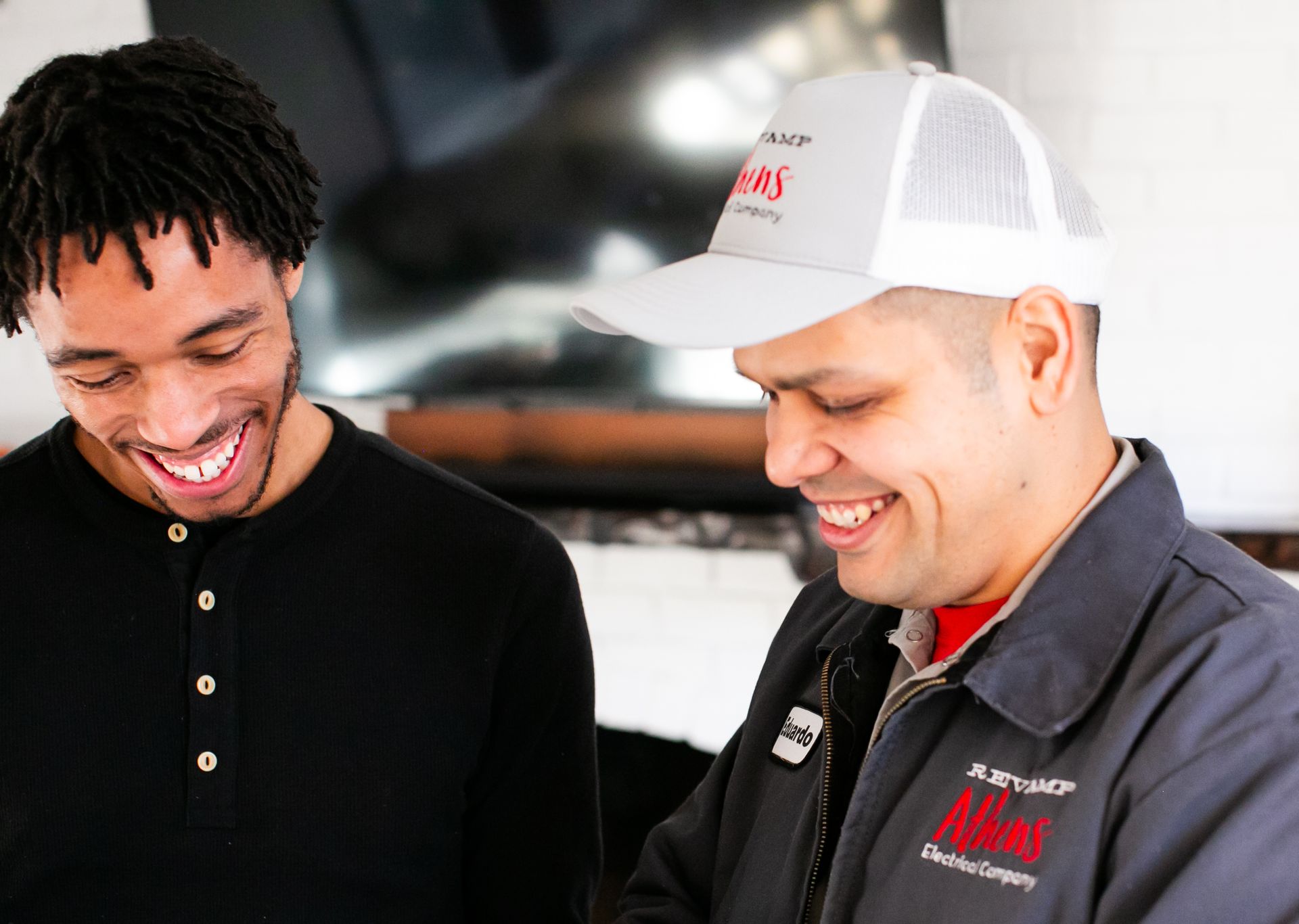 Two smiling workers in black shirts and caps looking at something together indoors
