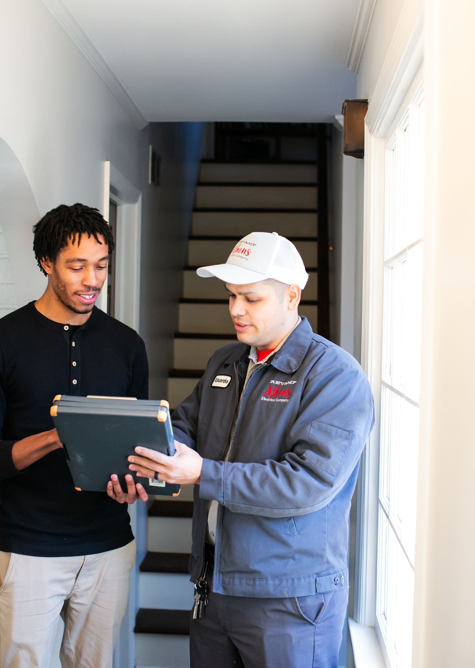 Two men reviewing a tablet in a narrow hallway beside a staircase.
