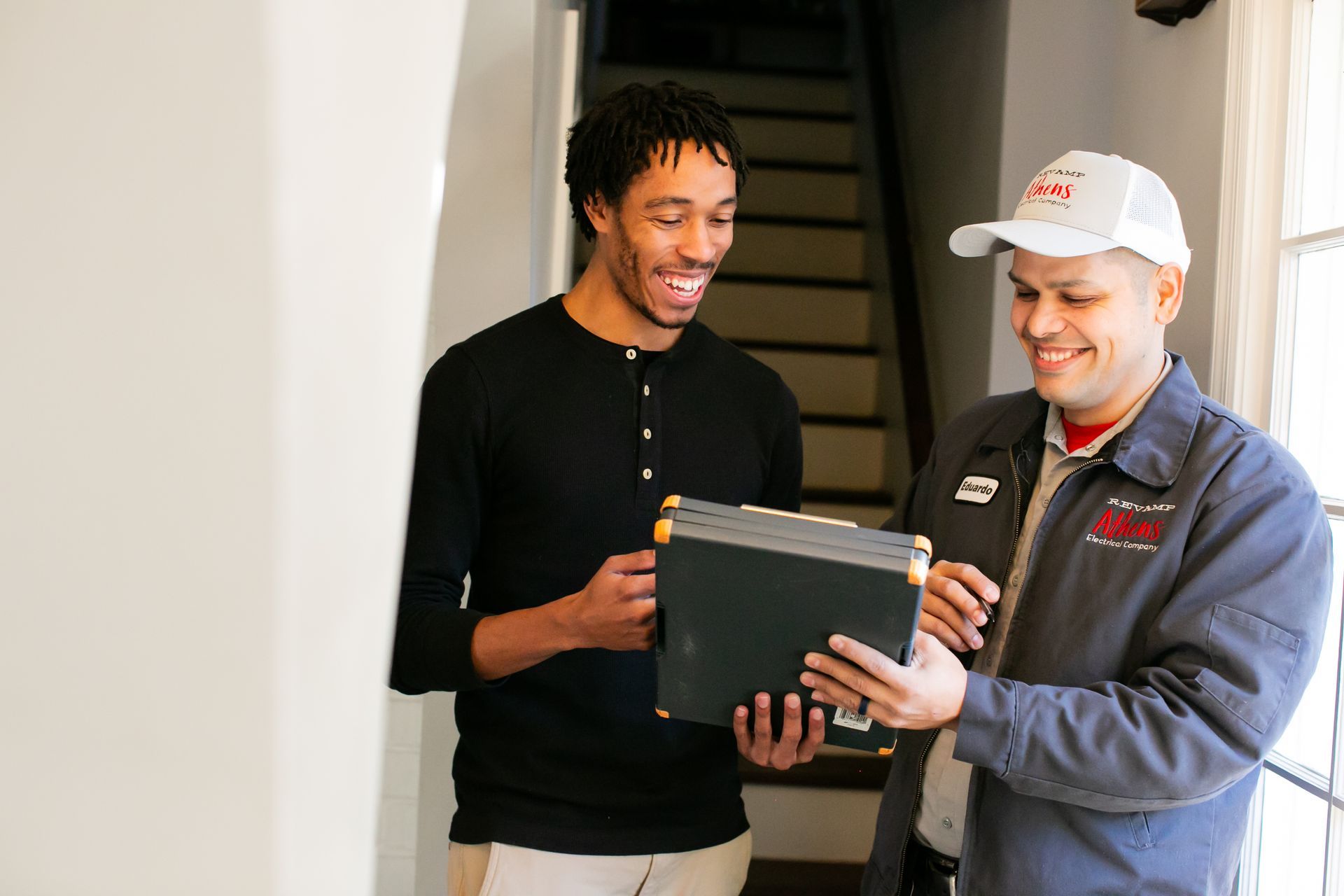 Two smiling people reviewing a laptop in a hallway with stairs behind them