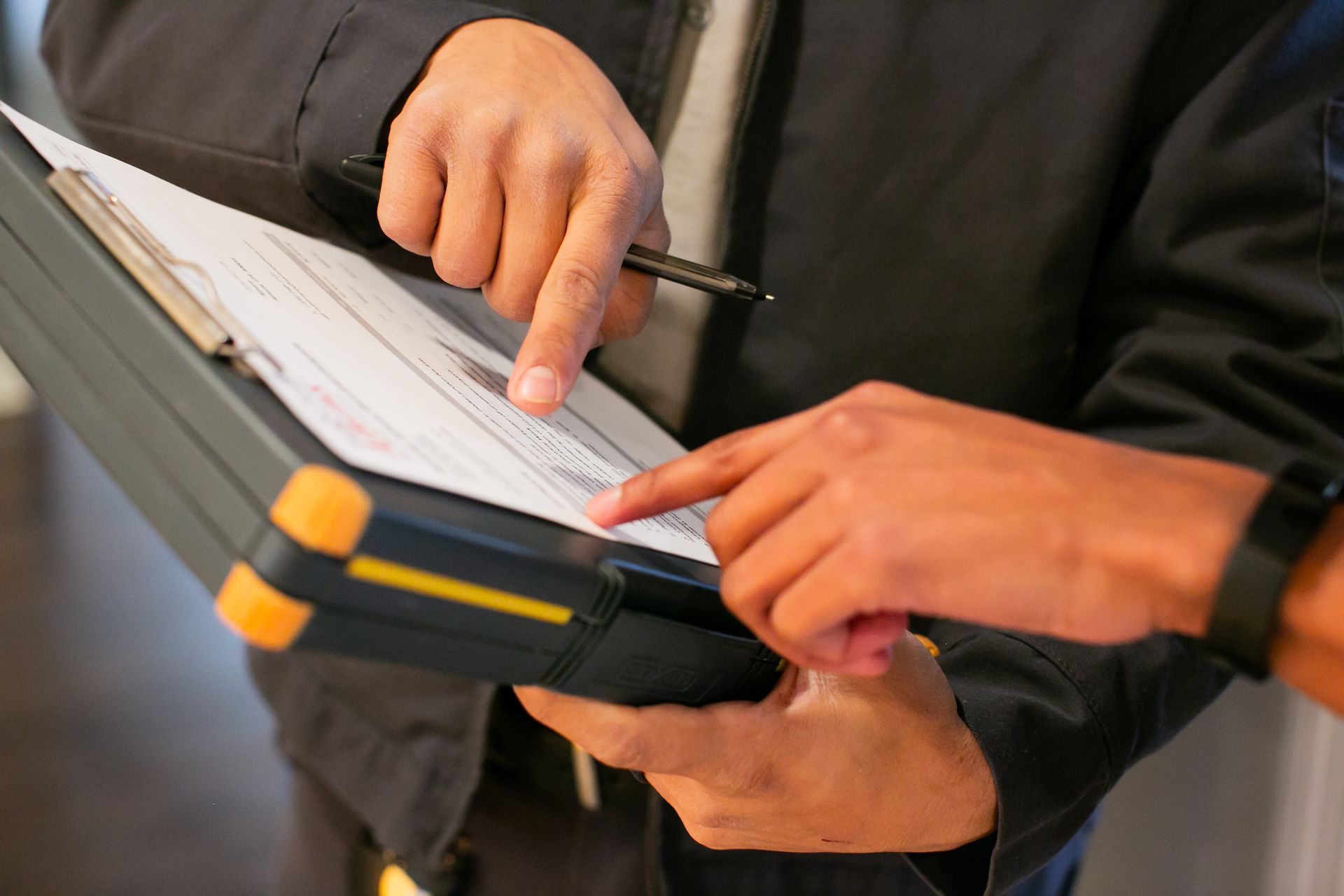 Hands writing on a clipboard with a pen, holding a tablet in a professional setting