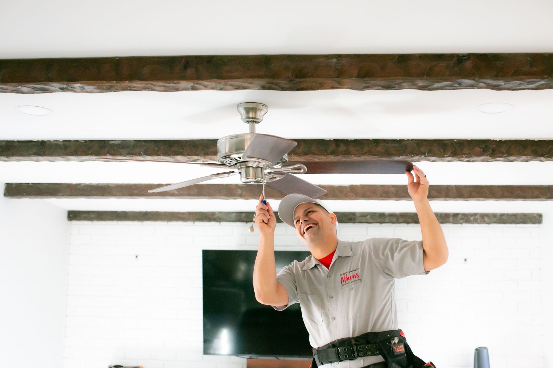 Technician adjusting a ceiling fan under exposed wooden beams in a bright room