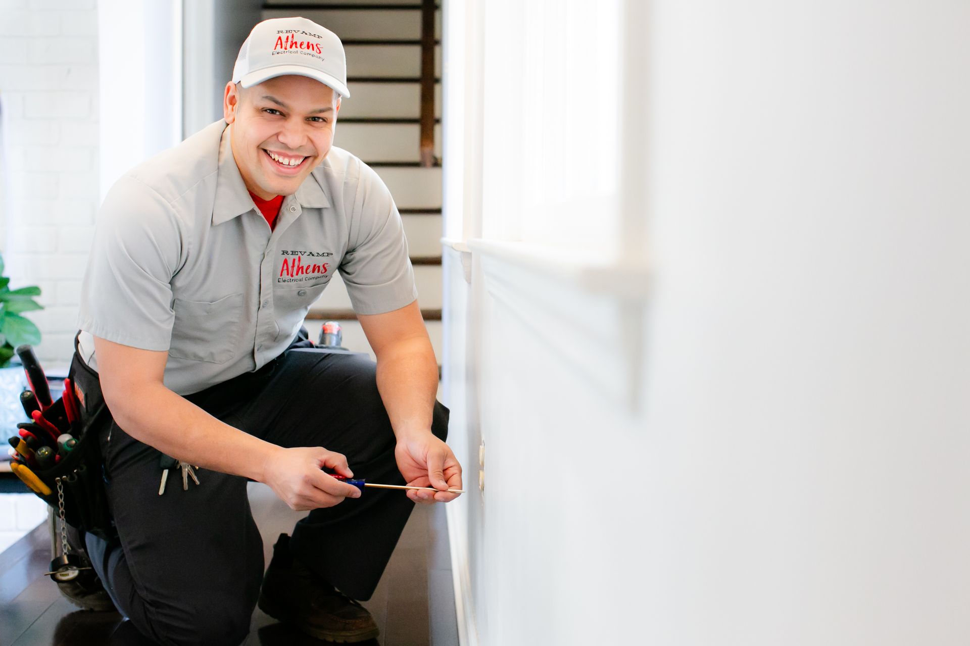 Smiling handyman kneels by a wall, holding a tool belt and wearing a gray uniform and cap.