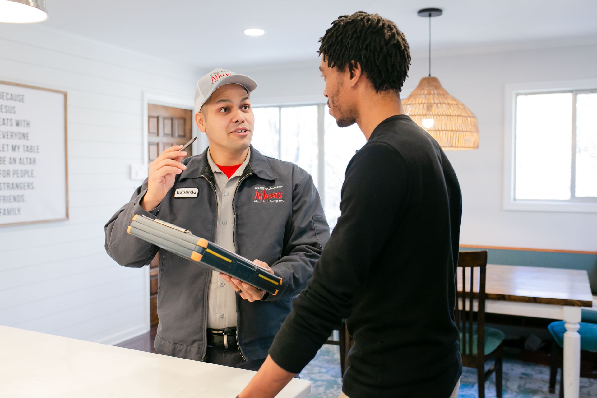 Two people talking in a bright home interior, one holding a tool or box on a kitchen counter.