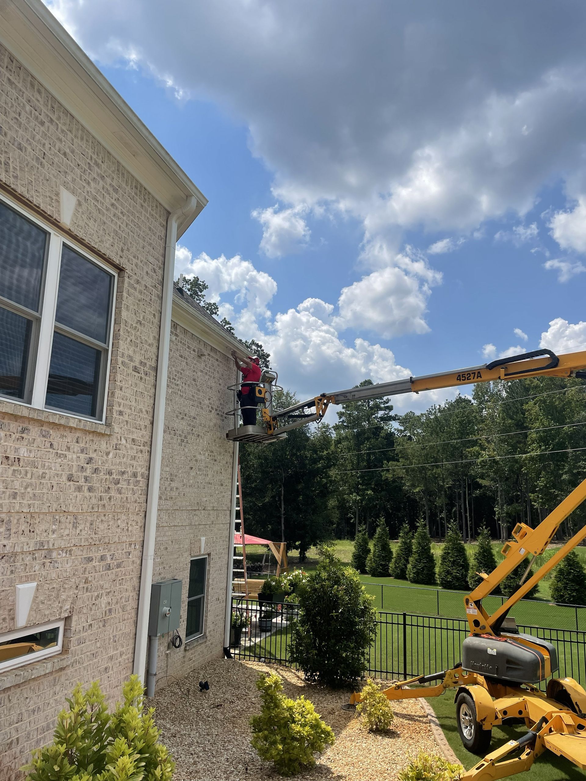 Worker on a ladder beside a stucco house, with a crane and trees in the backyard.