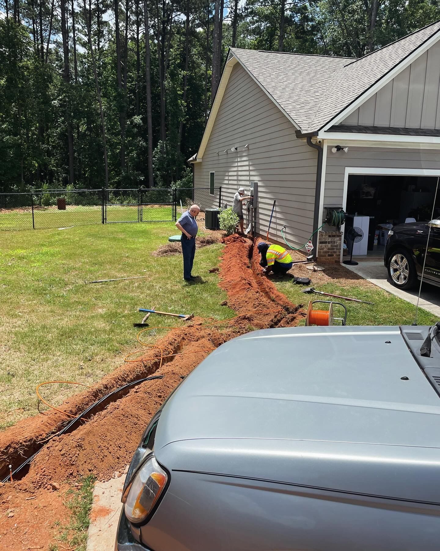 Workers digging a trench beside a gray house and garage, with a person standing nearby and a truck in front.
