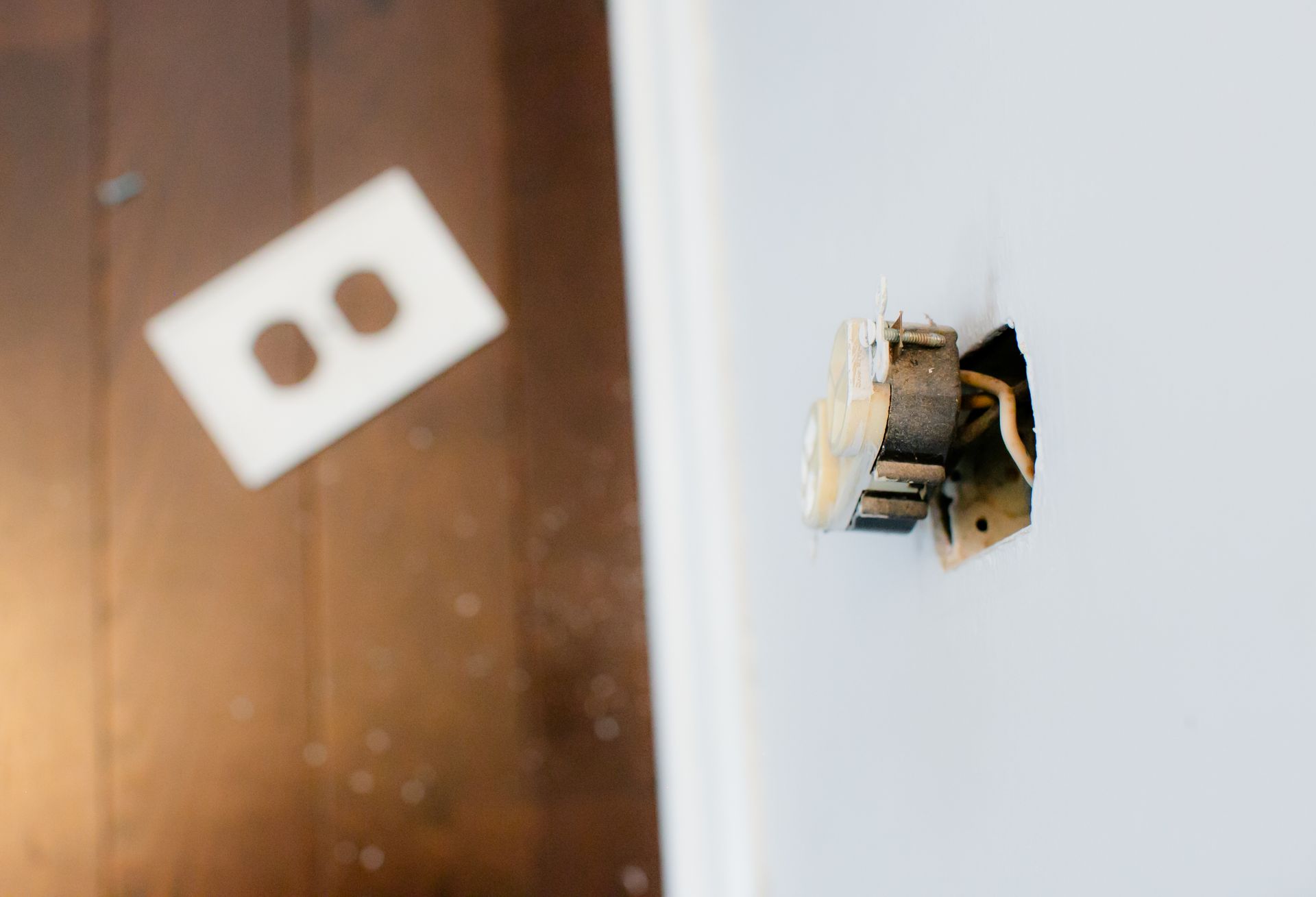 Close-up of a damaged wall outlet hole with exposed wires, beside a blurred white outlet cover.