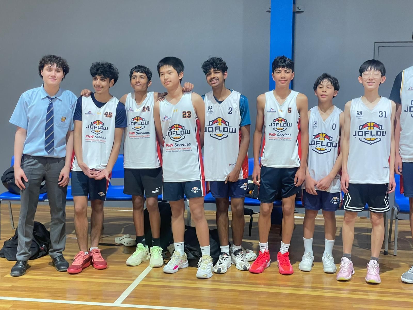 Basketball team of young boys posing for a photo in a gymnasium. Most wear jerseys; one in a school uniform.