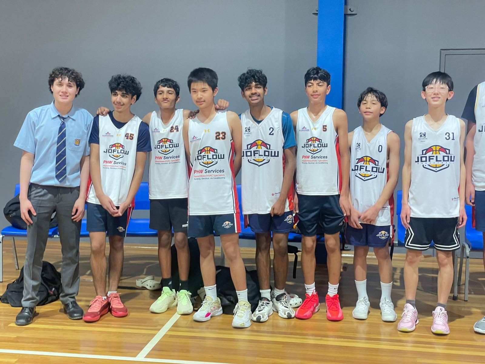 Youth basketball team poses on court. Players wear jerseys, one boy in school uniform.