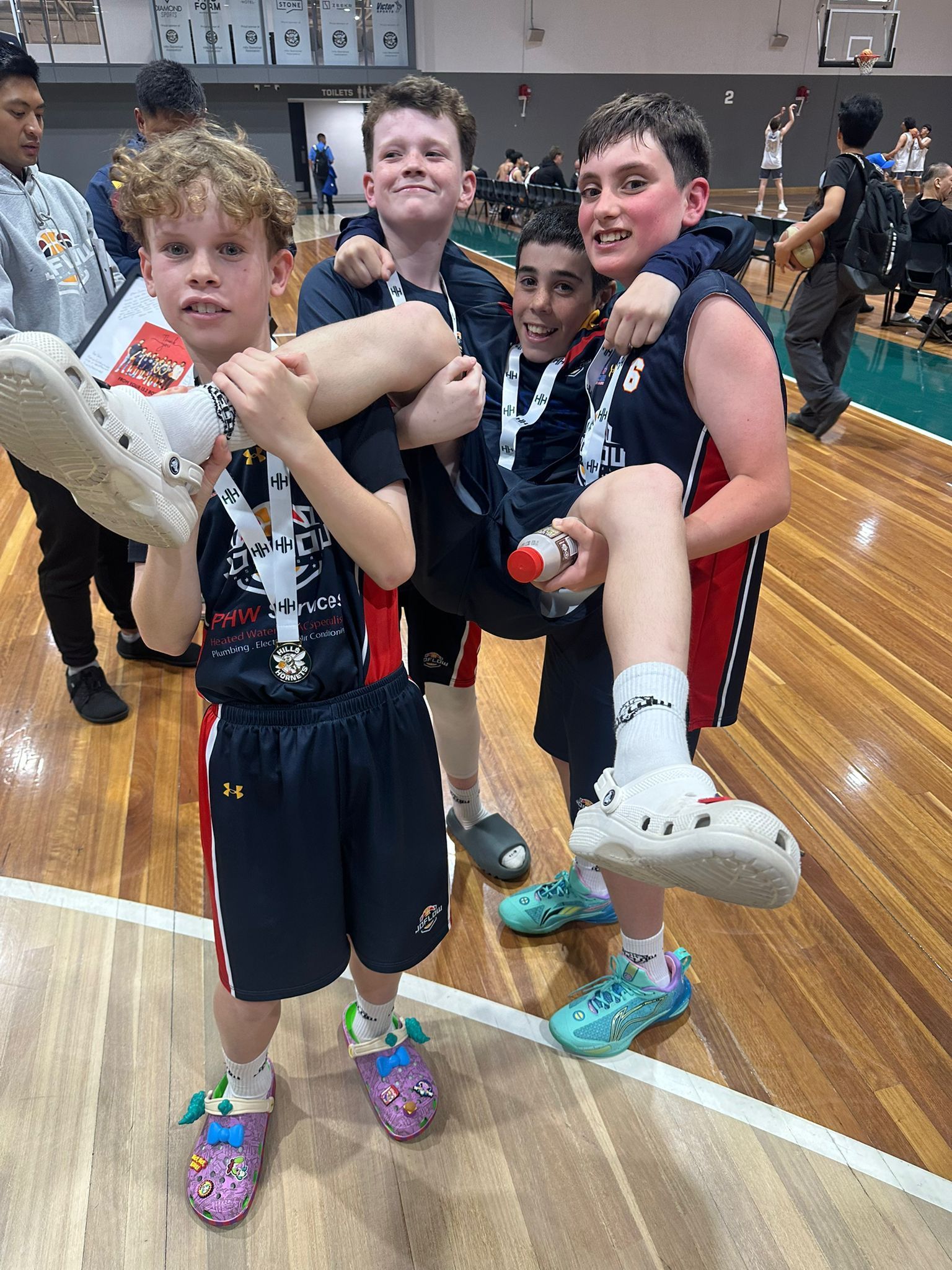 Basketball team celebrating win, two boys holding up another, wearing medals and Crocs on a court.