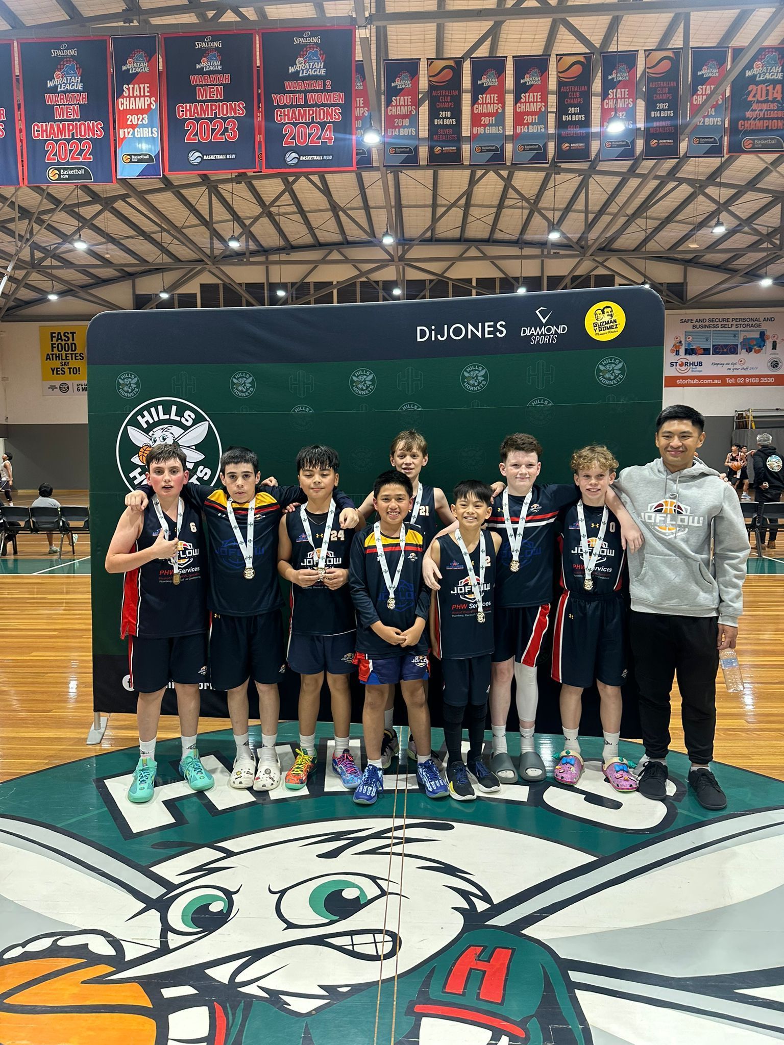 Youth basketball team posing with trophies on court, wearing medals. Green banner background.