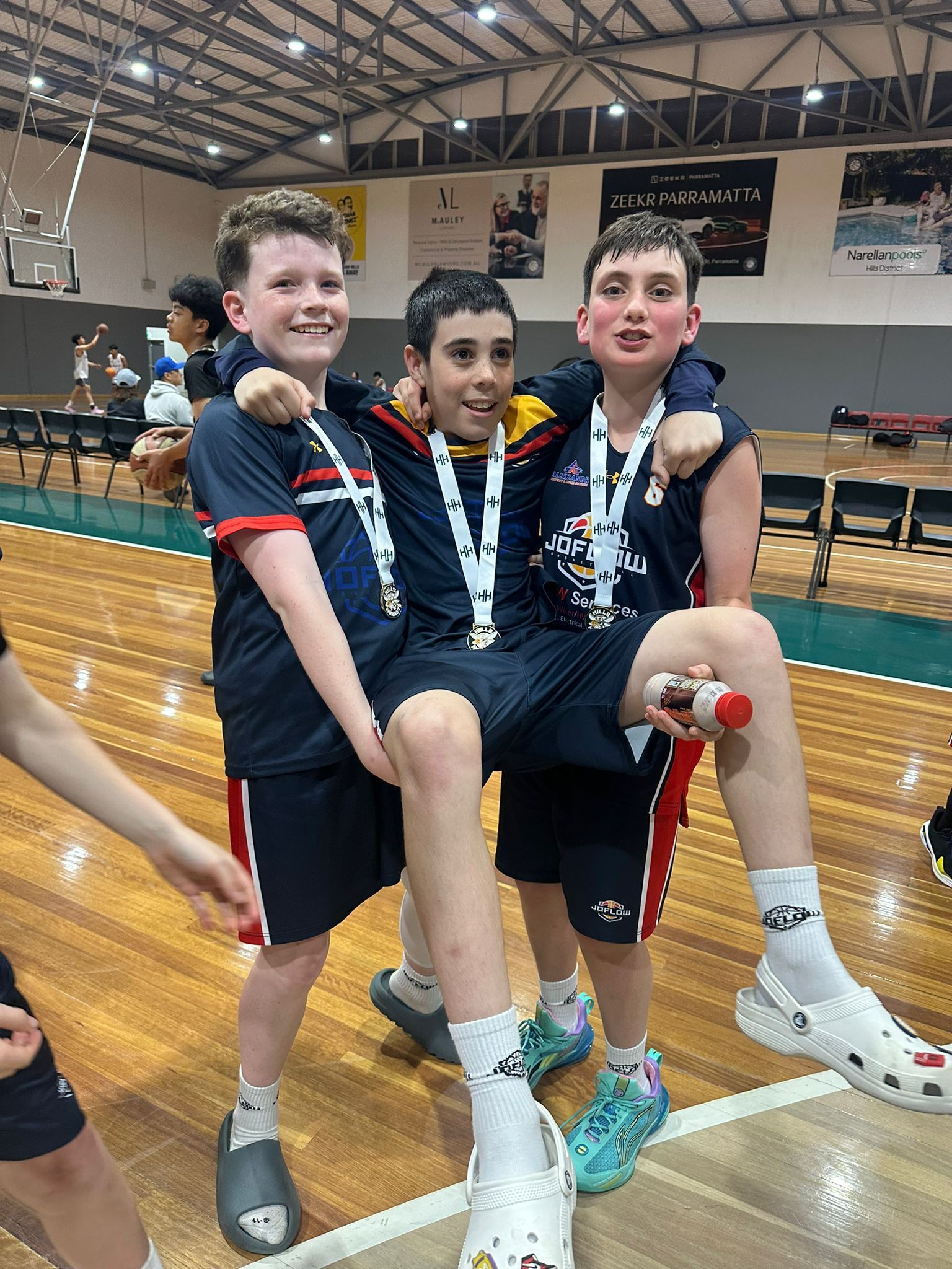 Three young basketball players, wearing medals, celebrating. They stand on a wooden court inside. One is being carried.