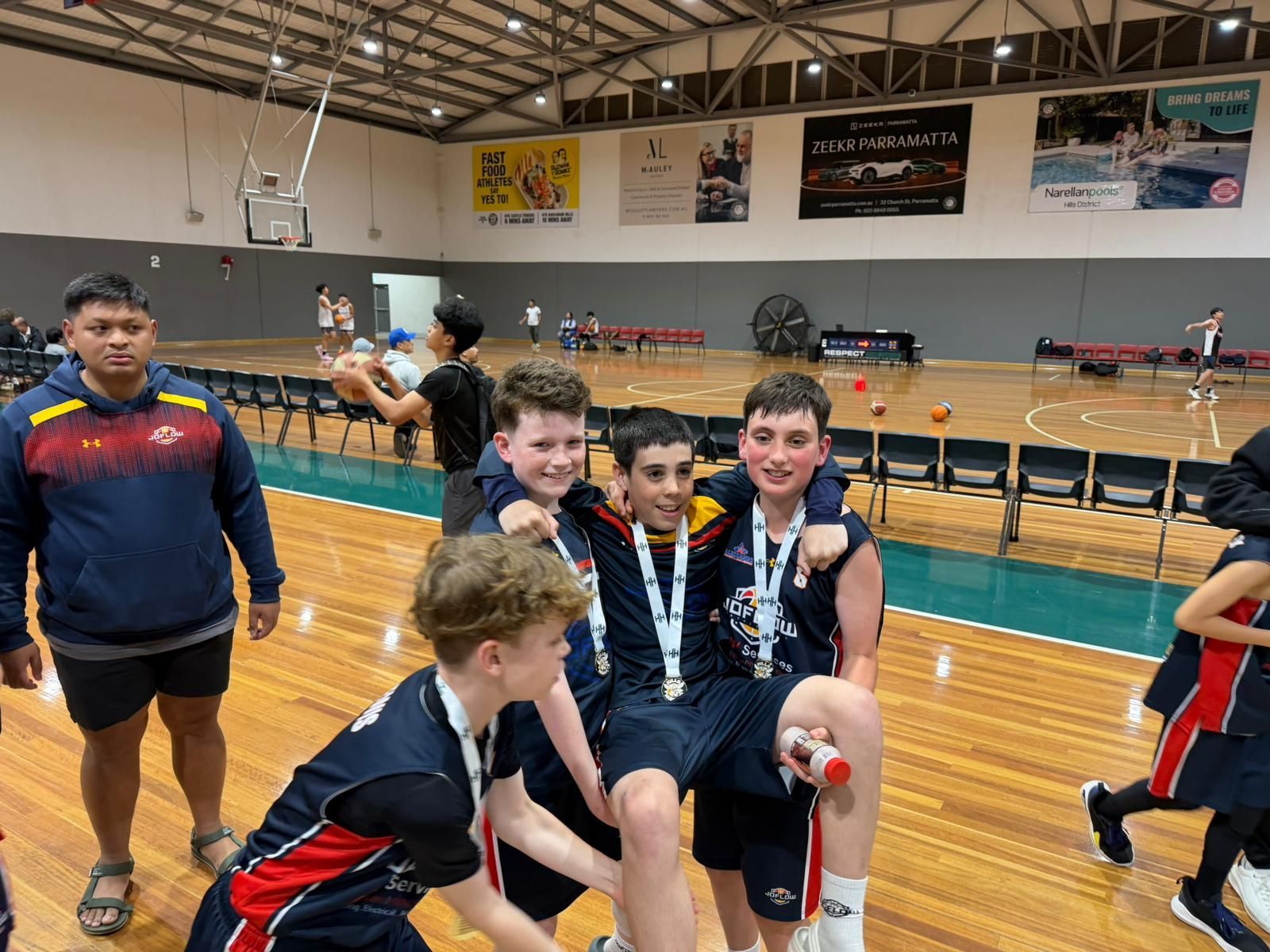 Basketball players celebrating with medals in a gymnasium; some are carrying a teammate, others look on.