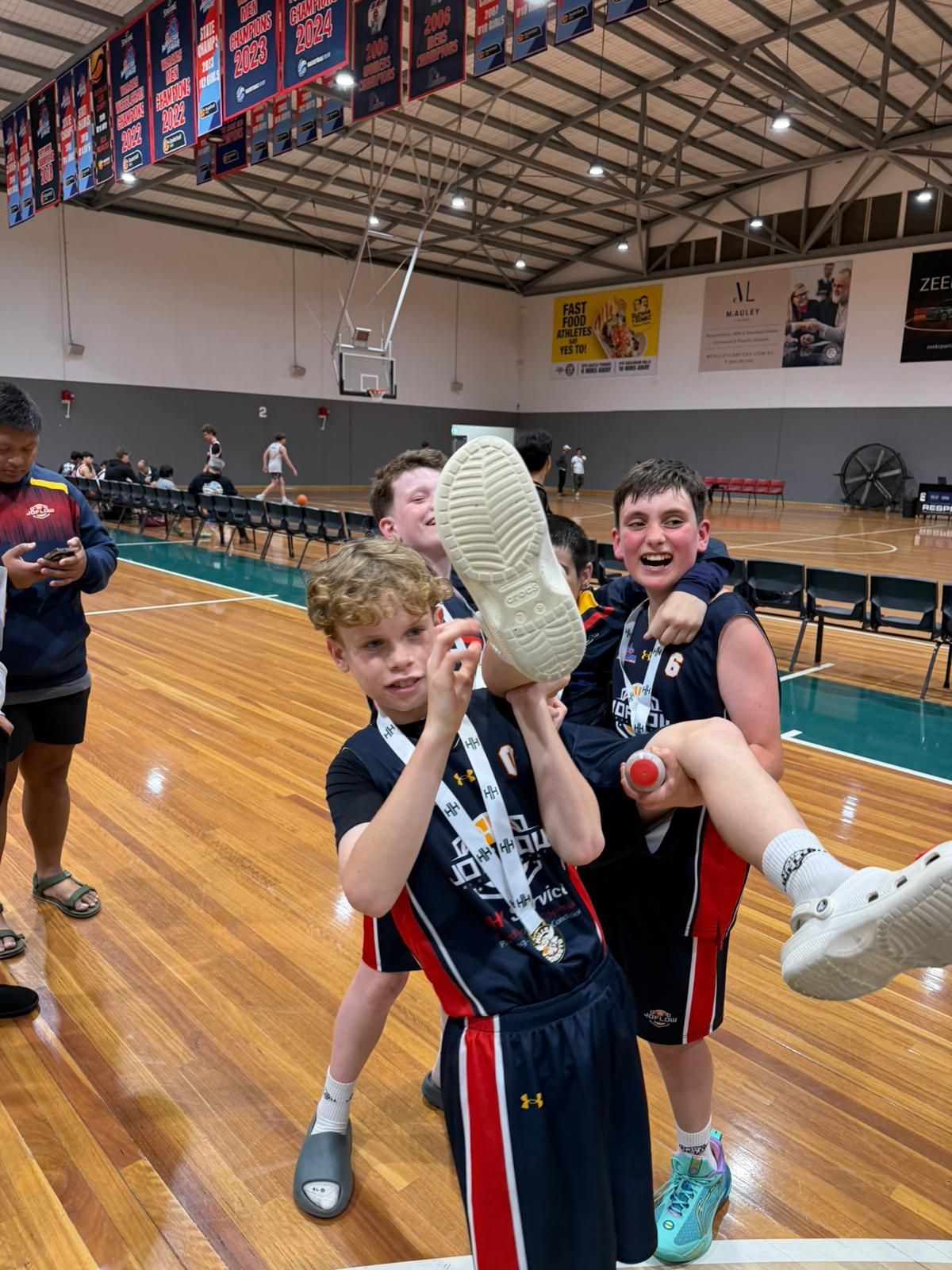 Boys celebrating on a basketball court, one holding a shoe up, all wearing medals, smiling.