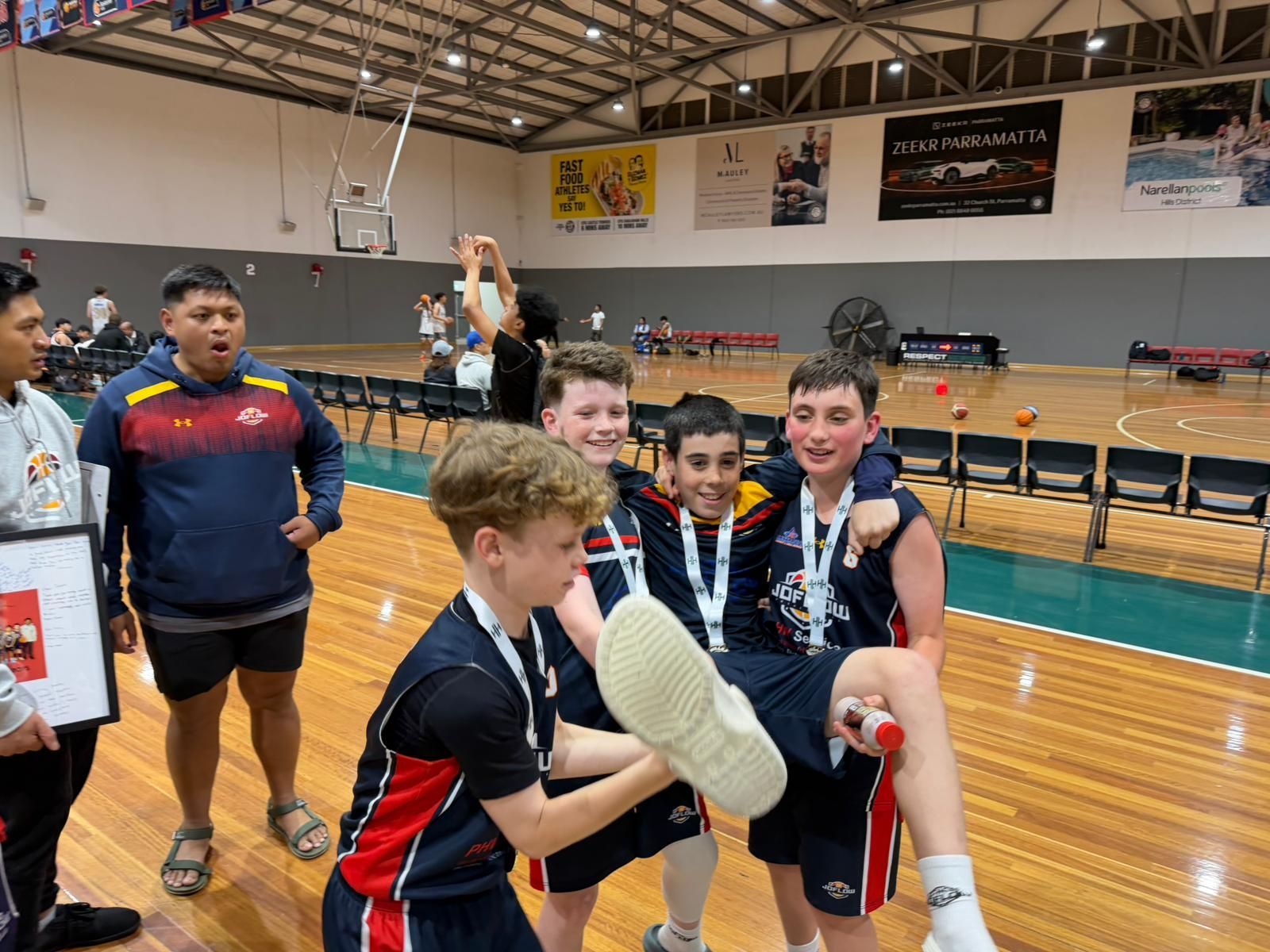Basketball team celebrating a win. Players lift a teammate while wearing medals. Indoor court.
