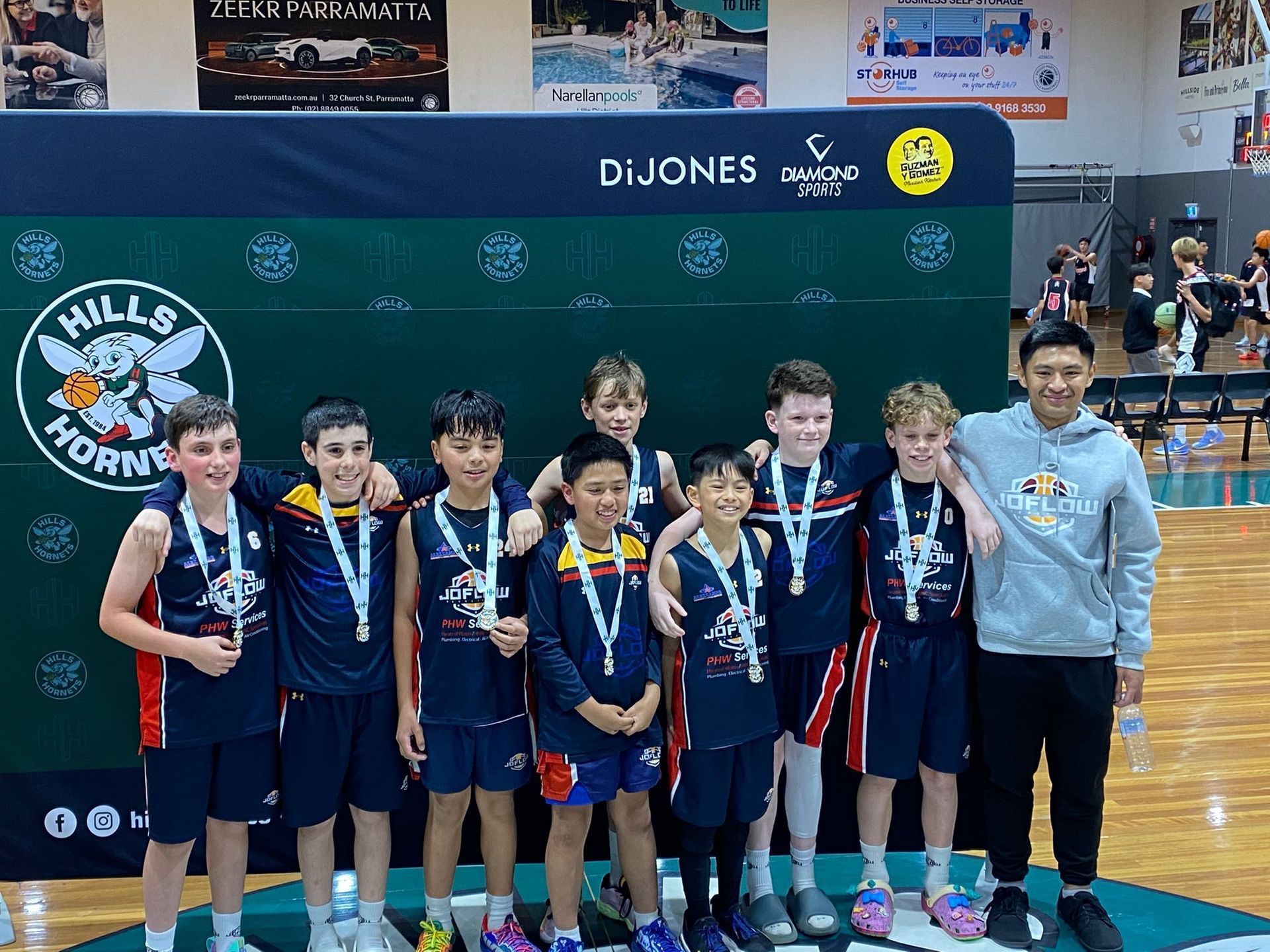Youth basketball team with medals posing after a game; indoor court, green background.