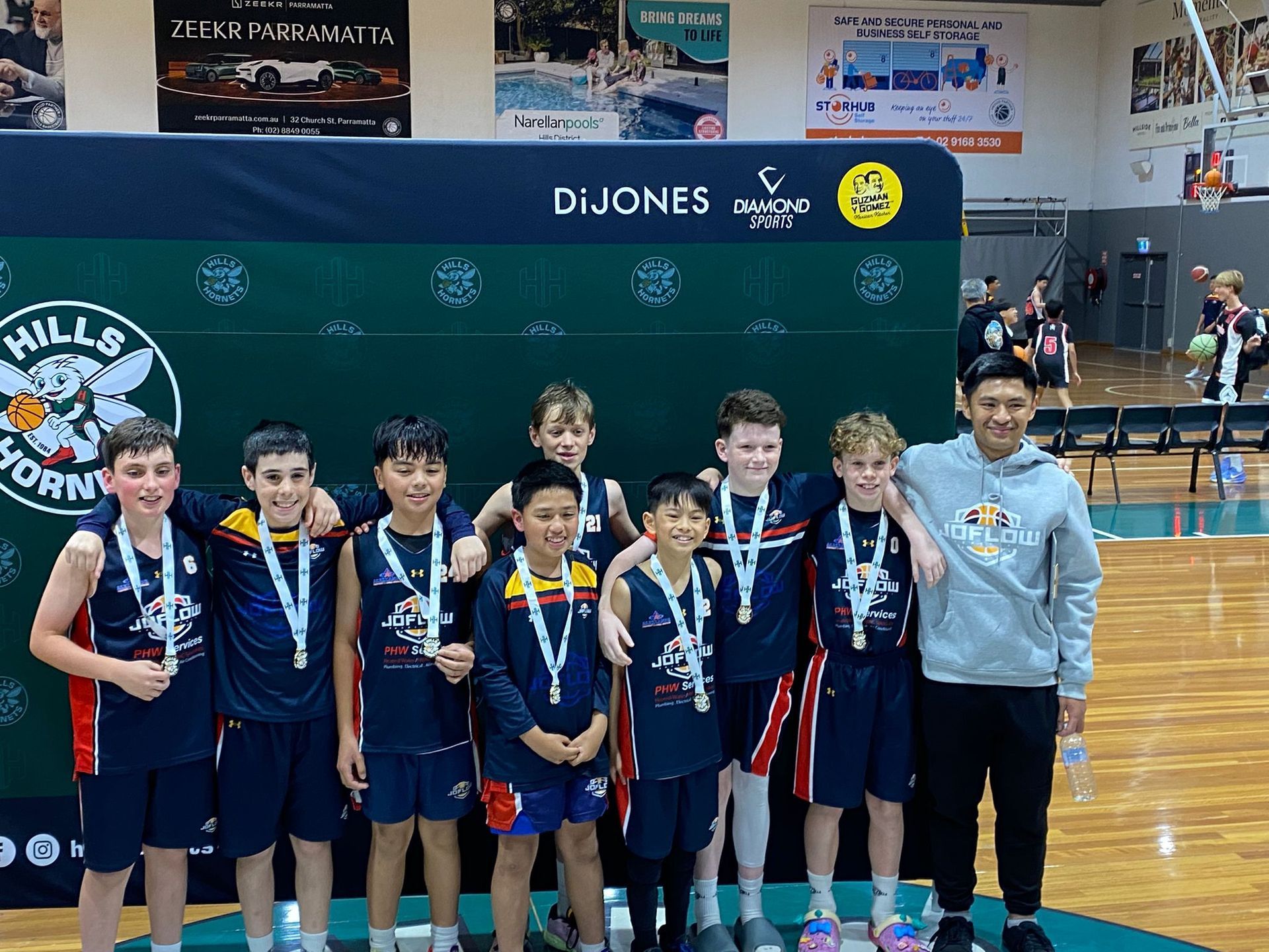 Basketball team with medals poses in front of a banner. Children and a coach smile.