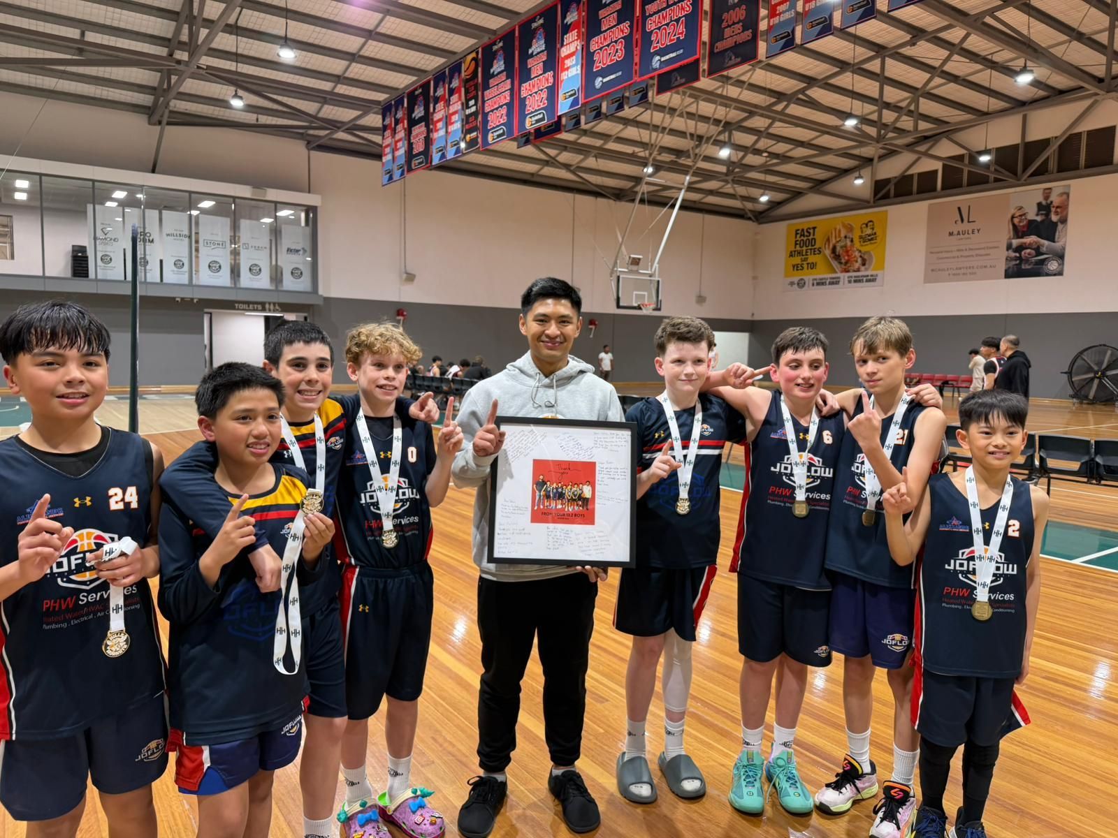 Youth basketball team poses with medals and a framed award in an indoor court.