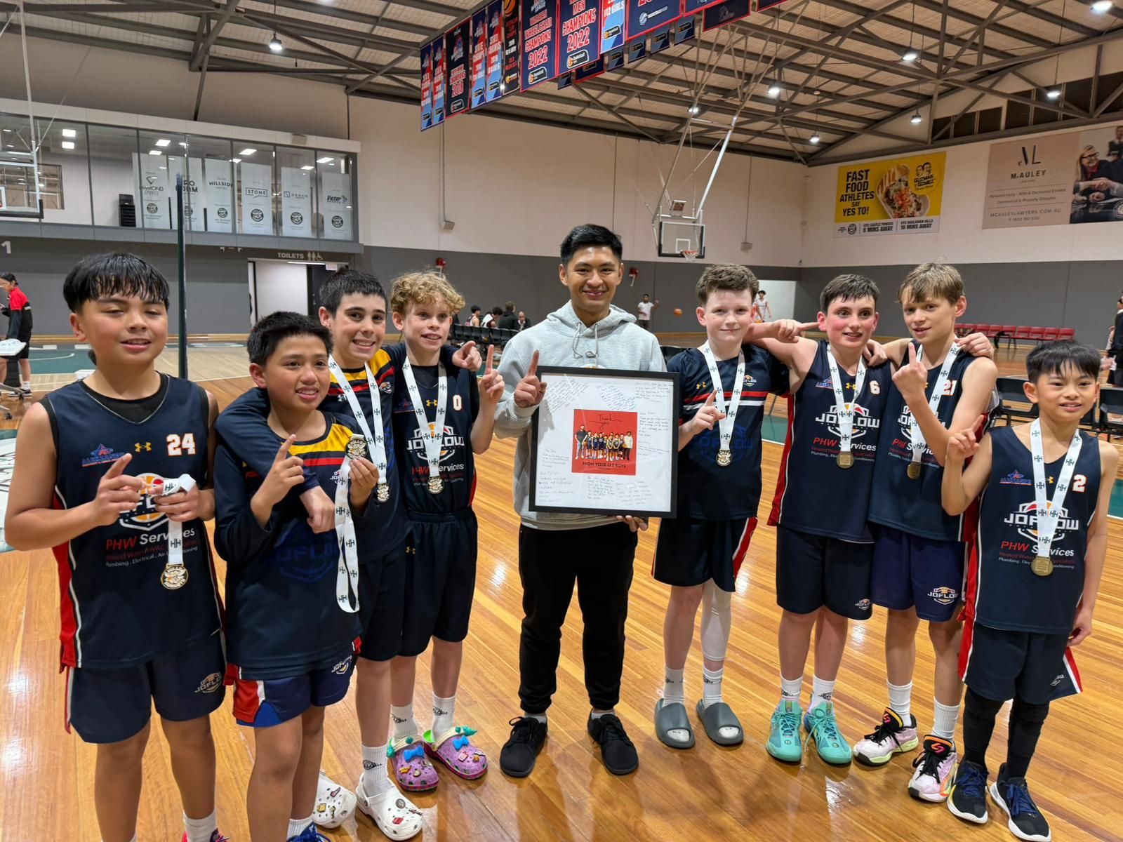 A basketball team poses with a framed photo, medals, and smiling. They are in a gymnasium.