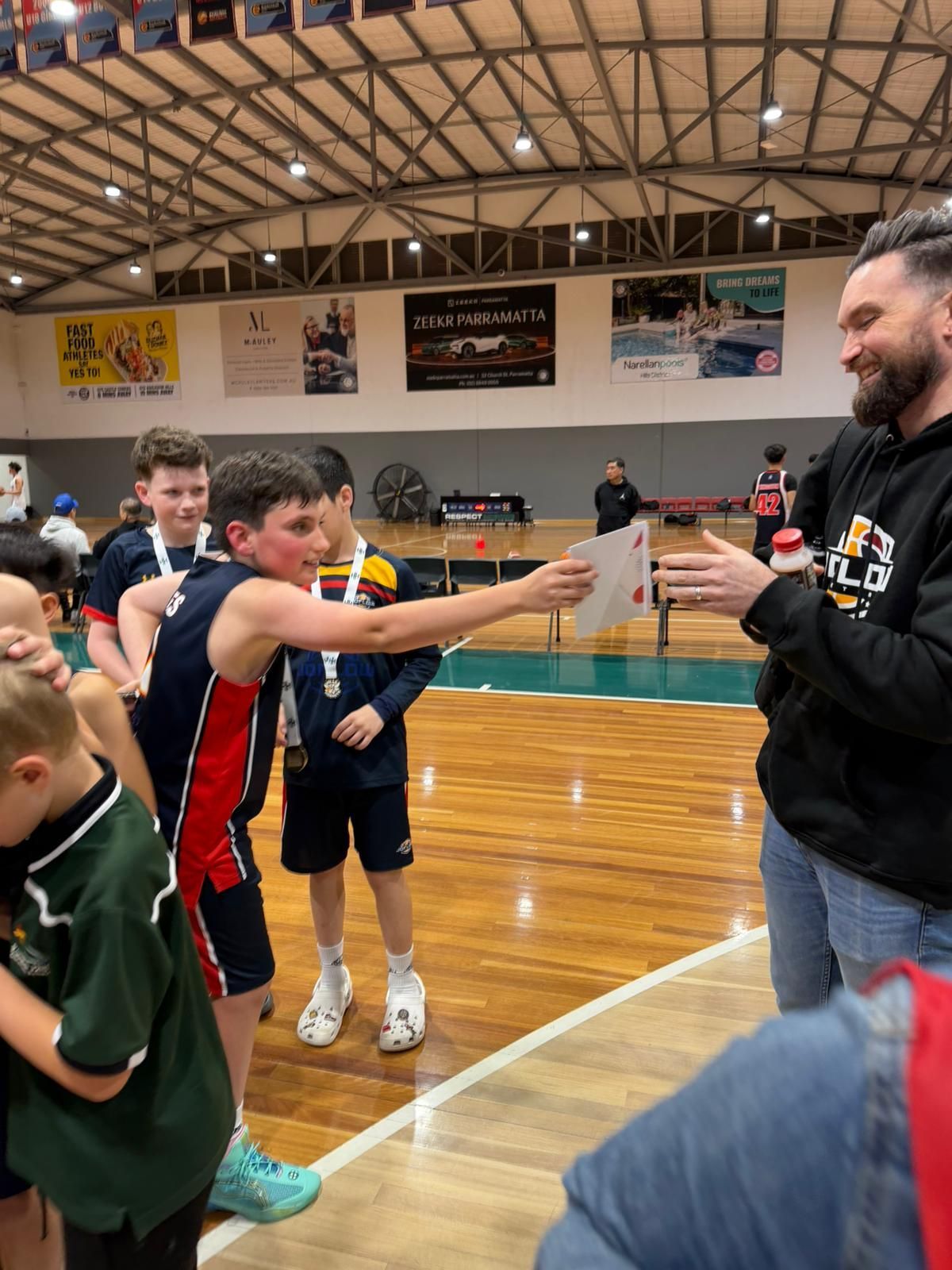 Boy in jersey handing a card to a man with a beard, indoor sports setting.